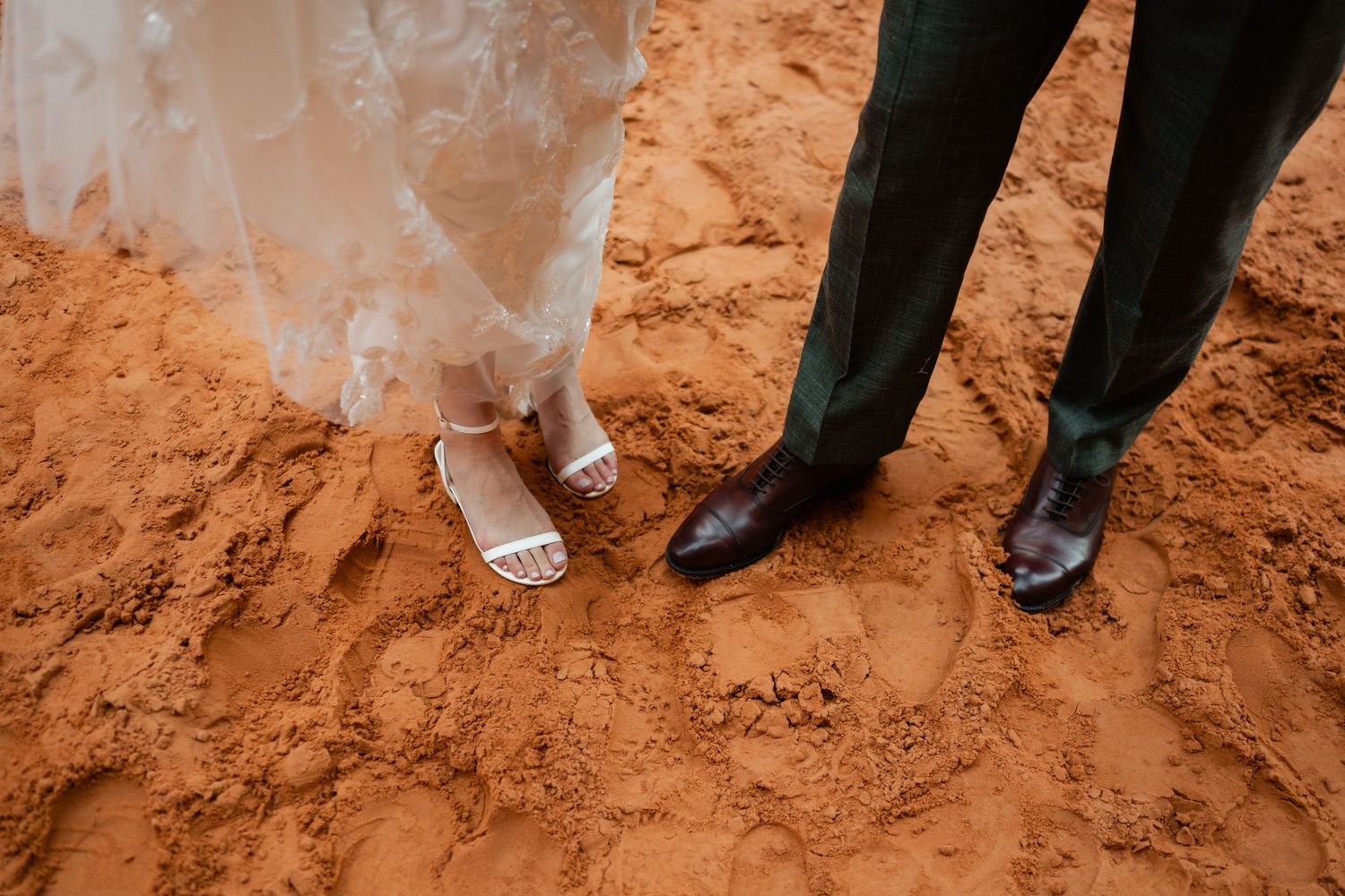 Close-up of formal shoes and white dress hem on red desert ground.