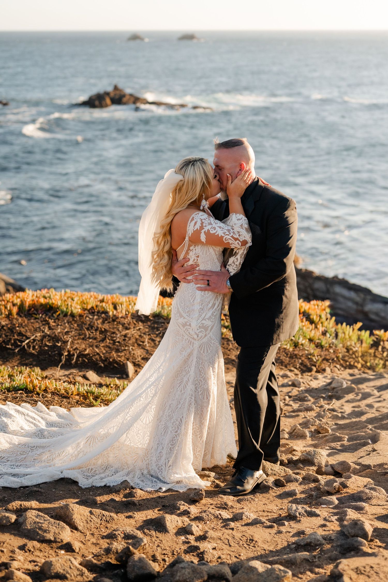 A romantic wedding couple embraces during golden hour on a rocky coastal cliff overlooking the ocean.