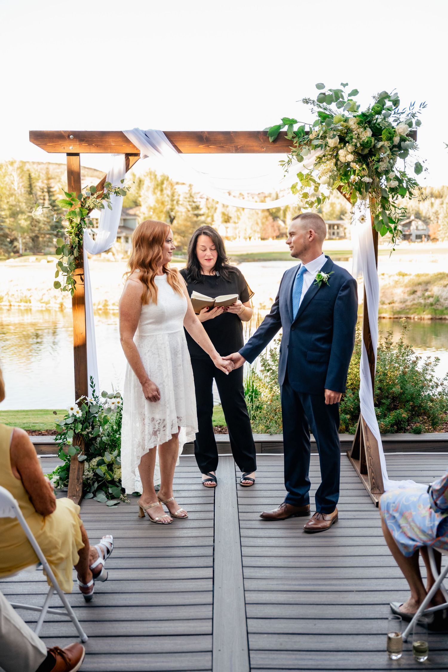 A wedding ceremony on a wooden deck by a lake shows feet and formal attire of attendees during an intimate gathering.