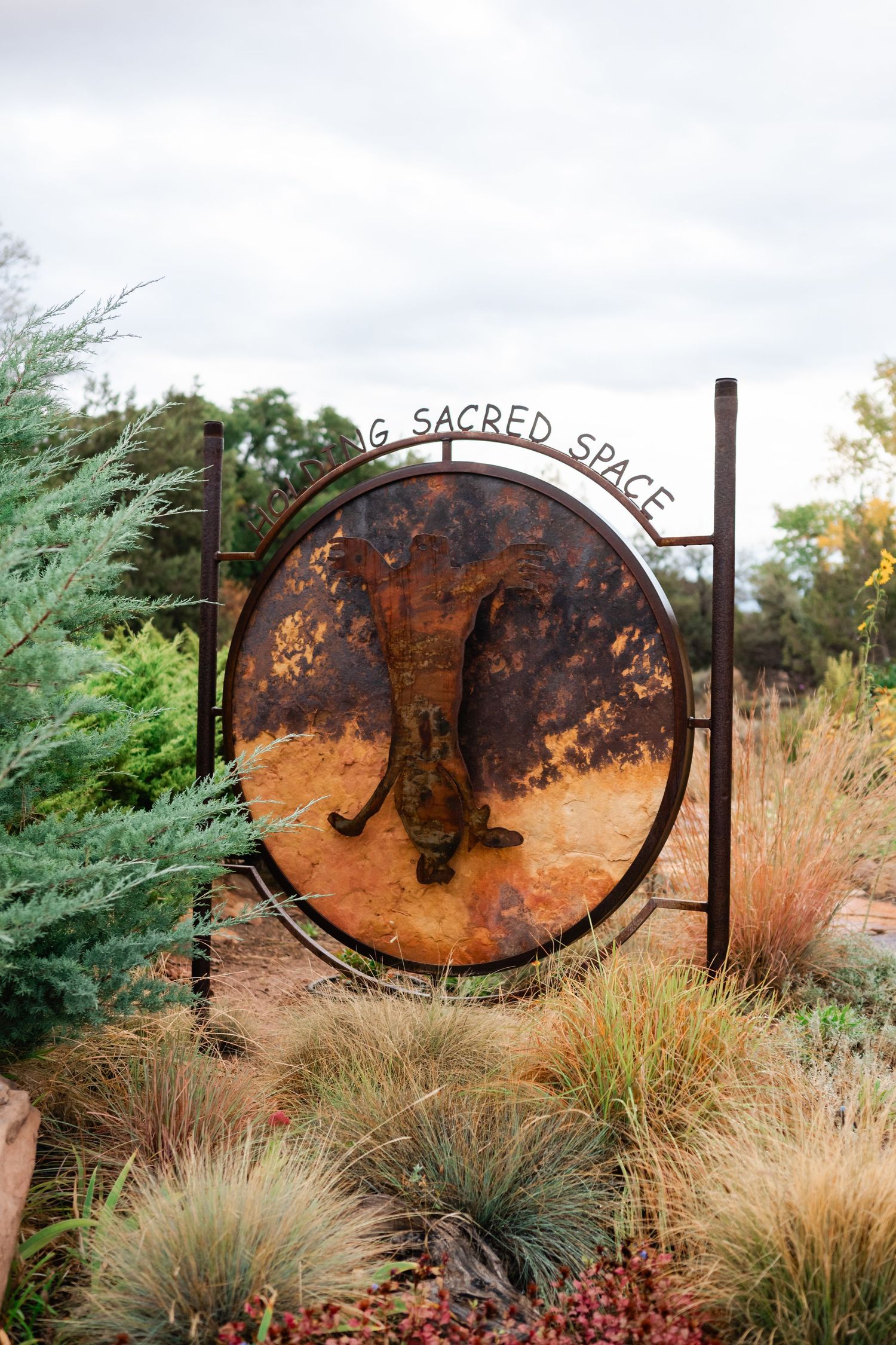 Rustic metal garden art featuring circular designs and text displayed among ornamental grasses in a landscaped setting.