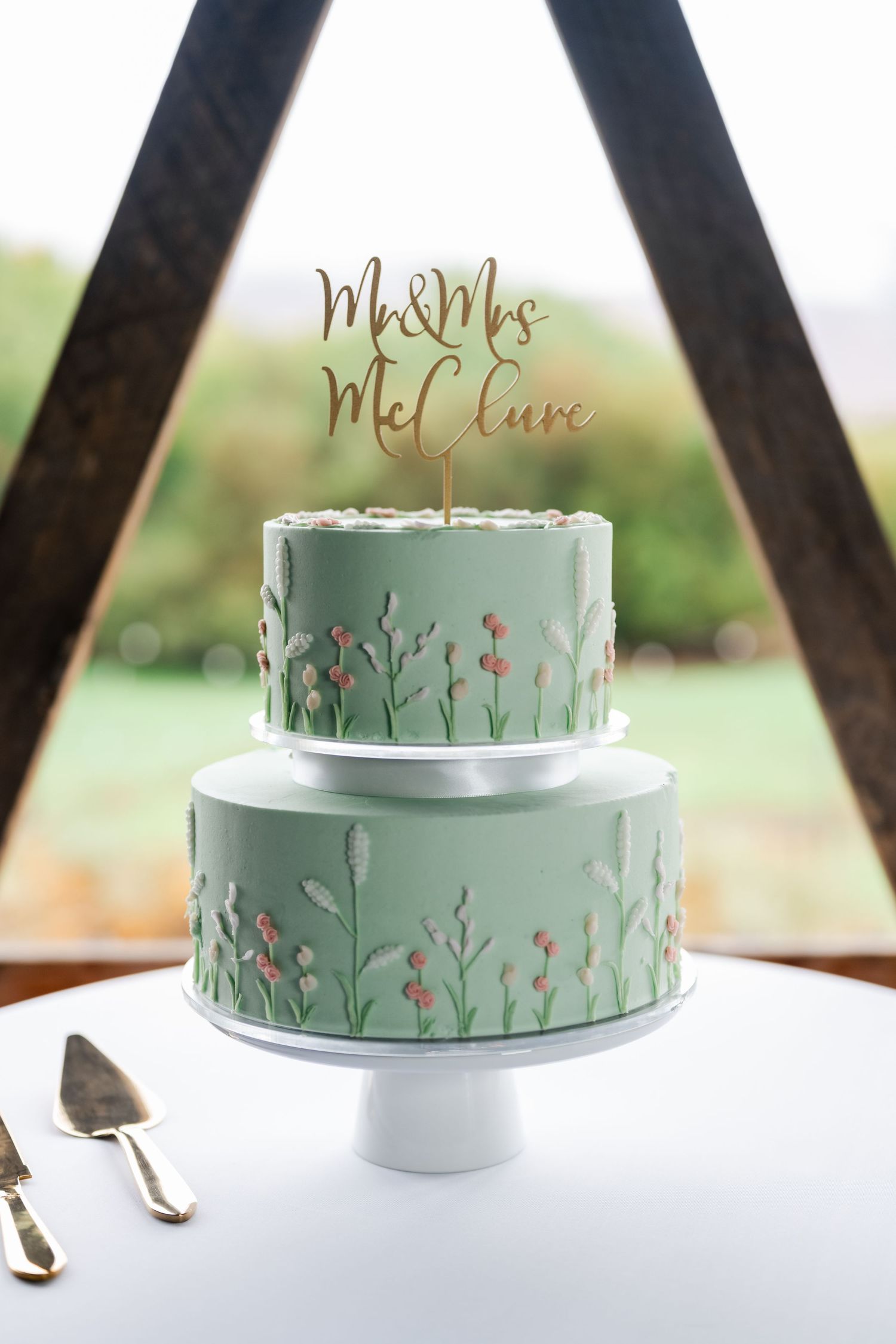 Two-tiered mint green wedding cake decorated with delicate floral designs and metallic name topper.