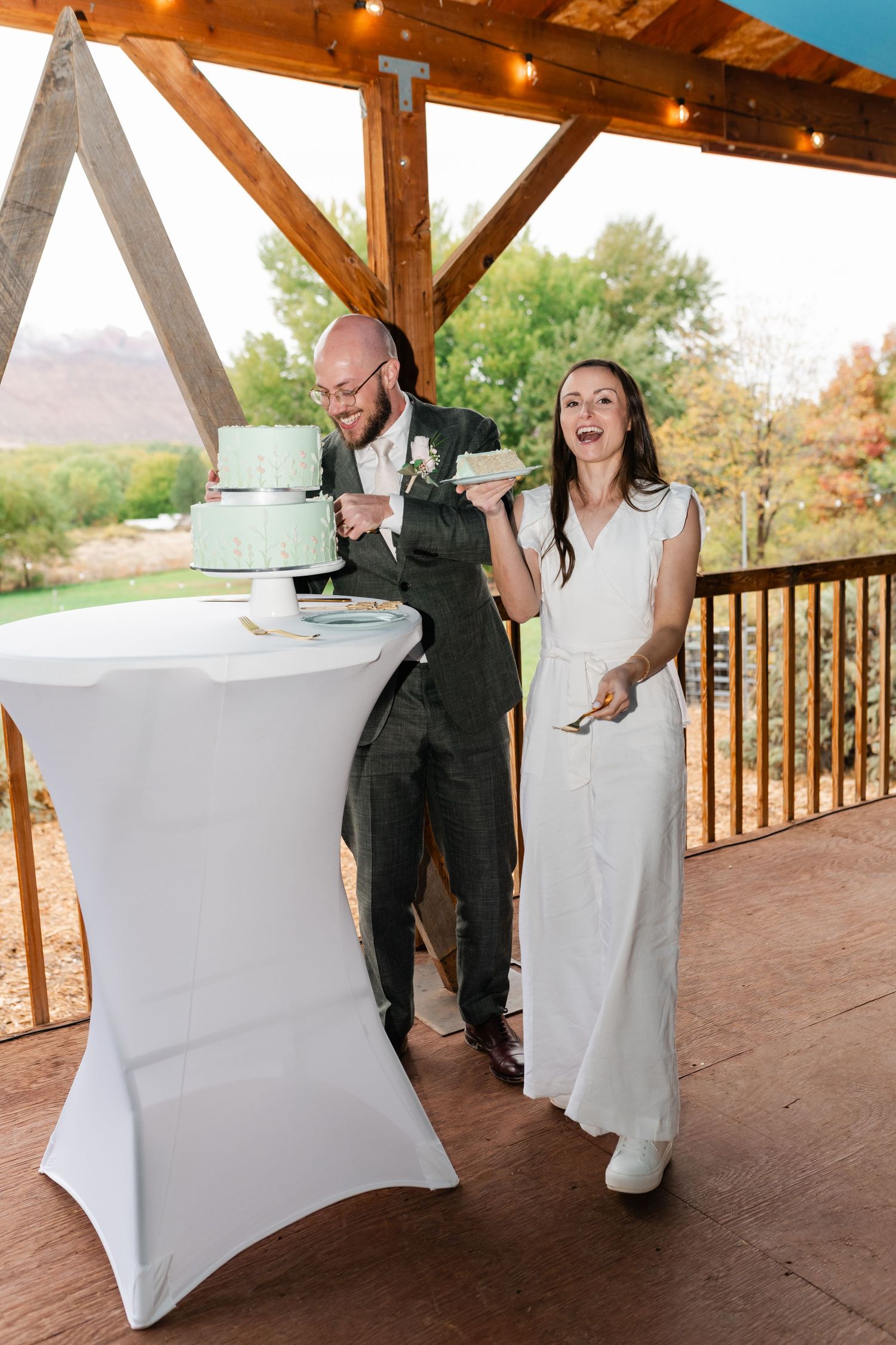 A couple sharing a joyful moment while cutting their mint green wedding cake on a covered patio.