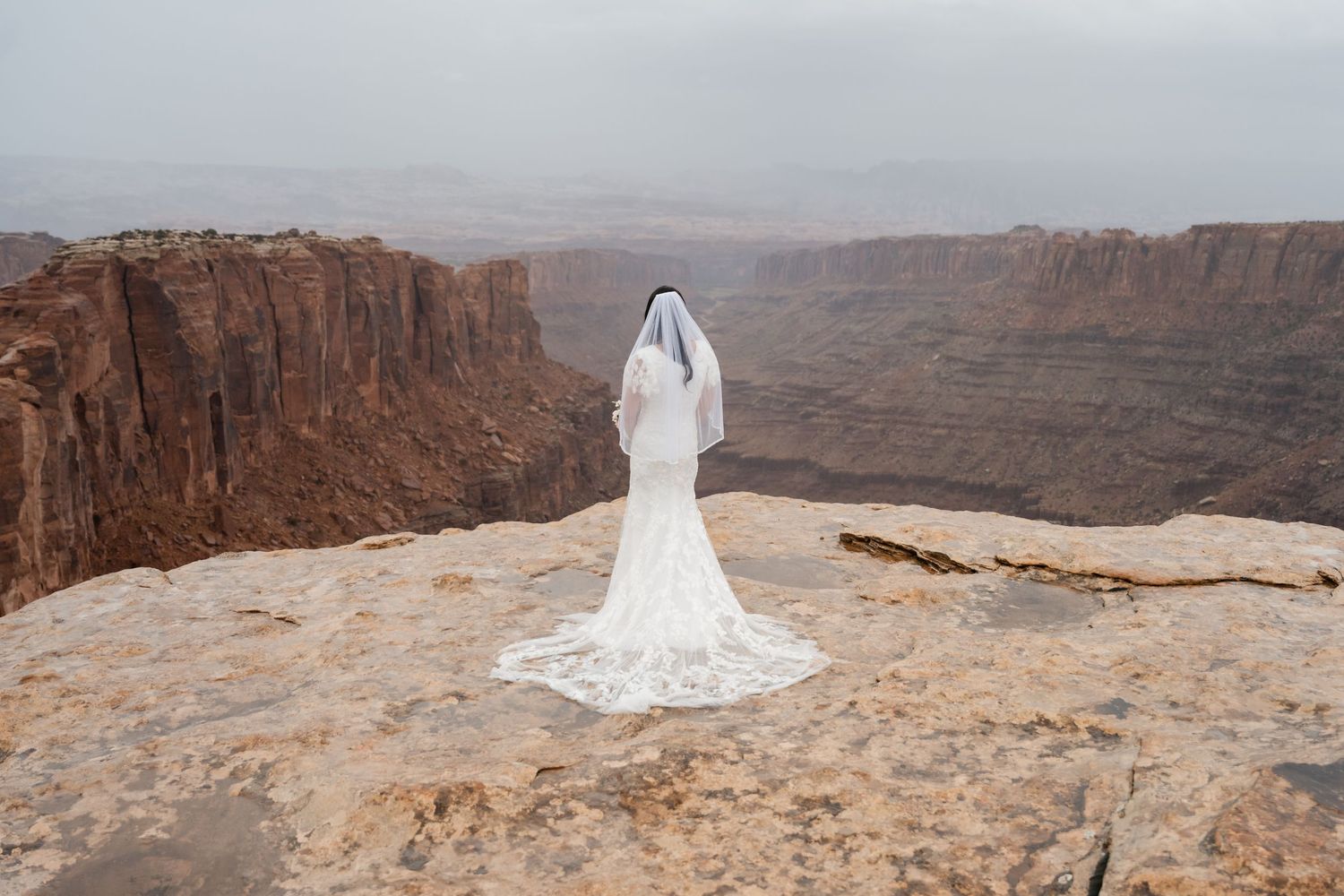A long flowing white wedding dress trails across rugged desert cliffs with sweeping canyon vistas in the distance.