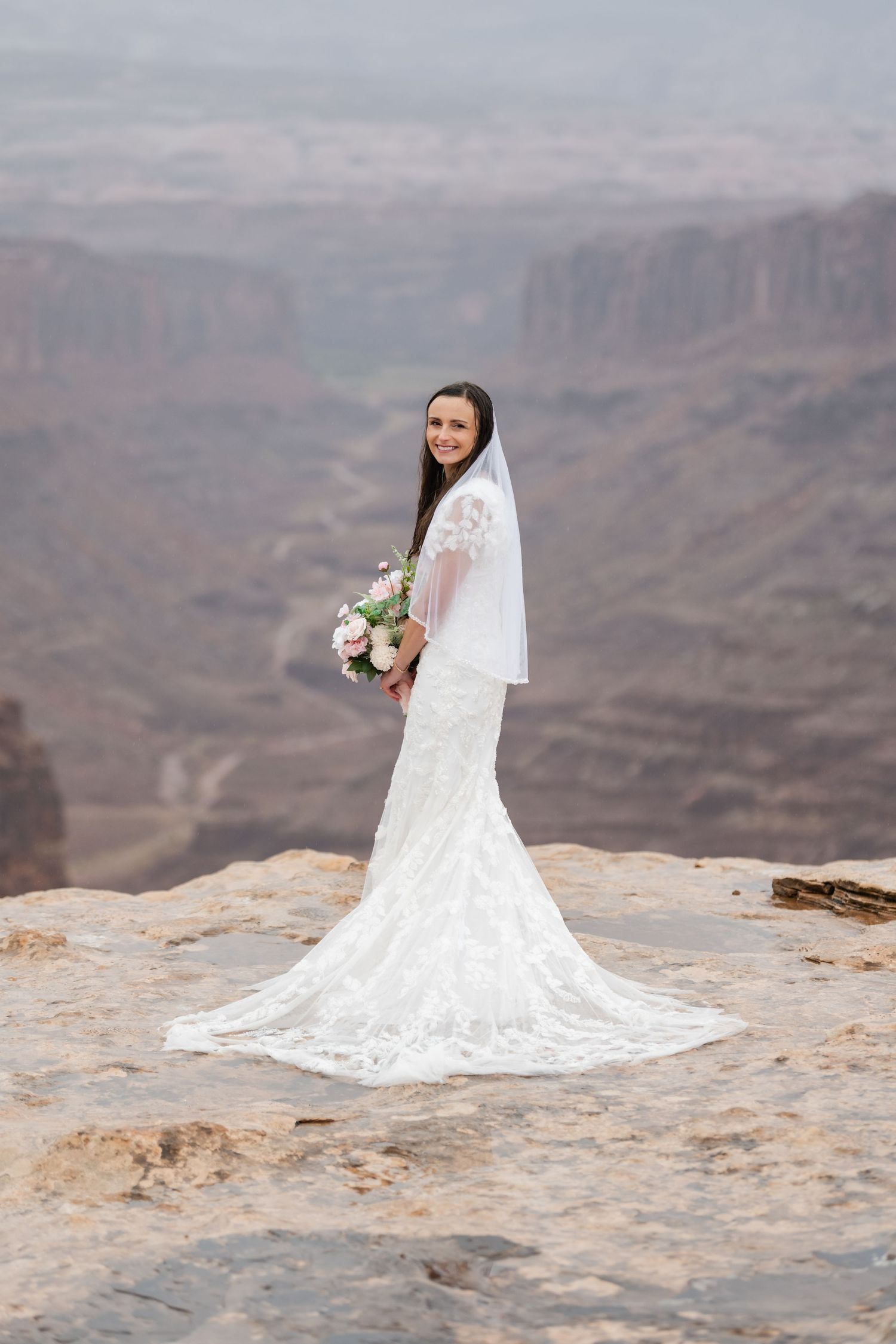 A bride in a flowing lace dress poses against a stunning canyon backdrop.