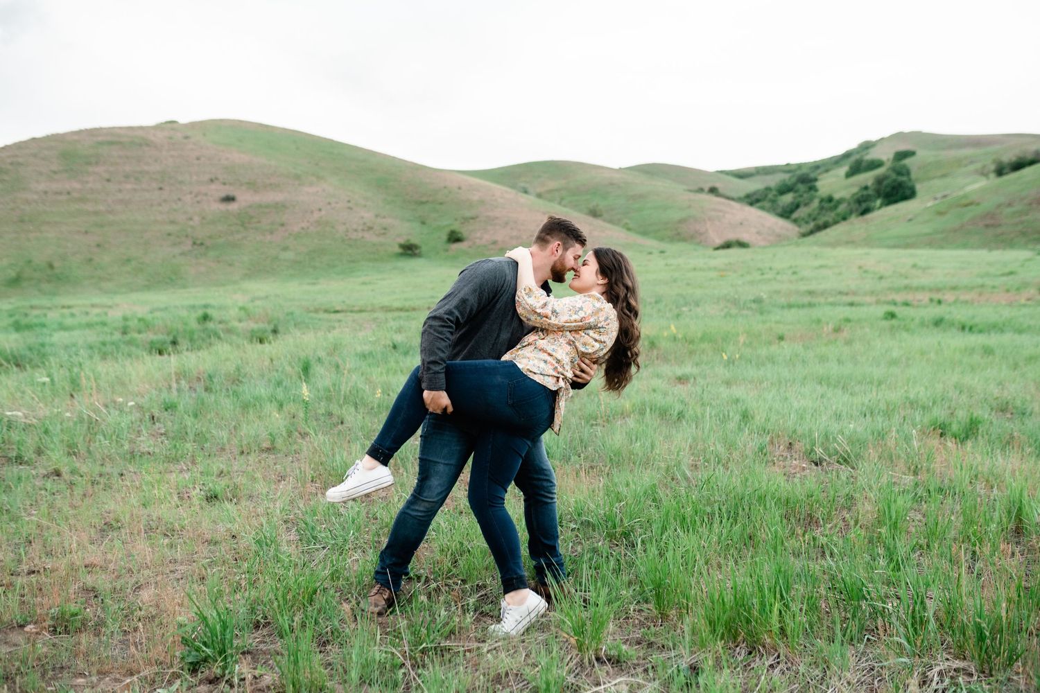A romantic couple embraces in a scenic green meadow with rolling hills in the background during a photo session.