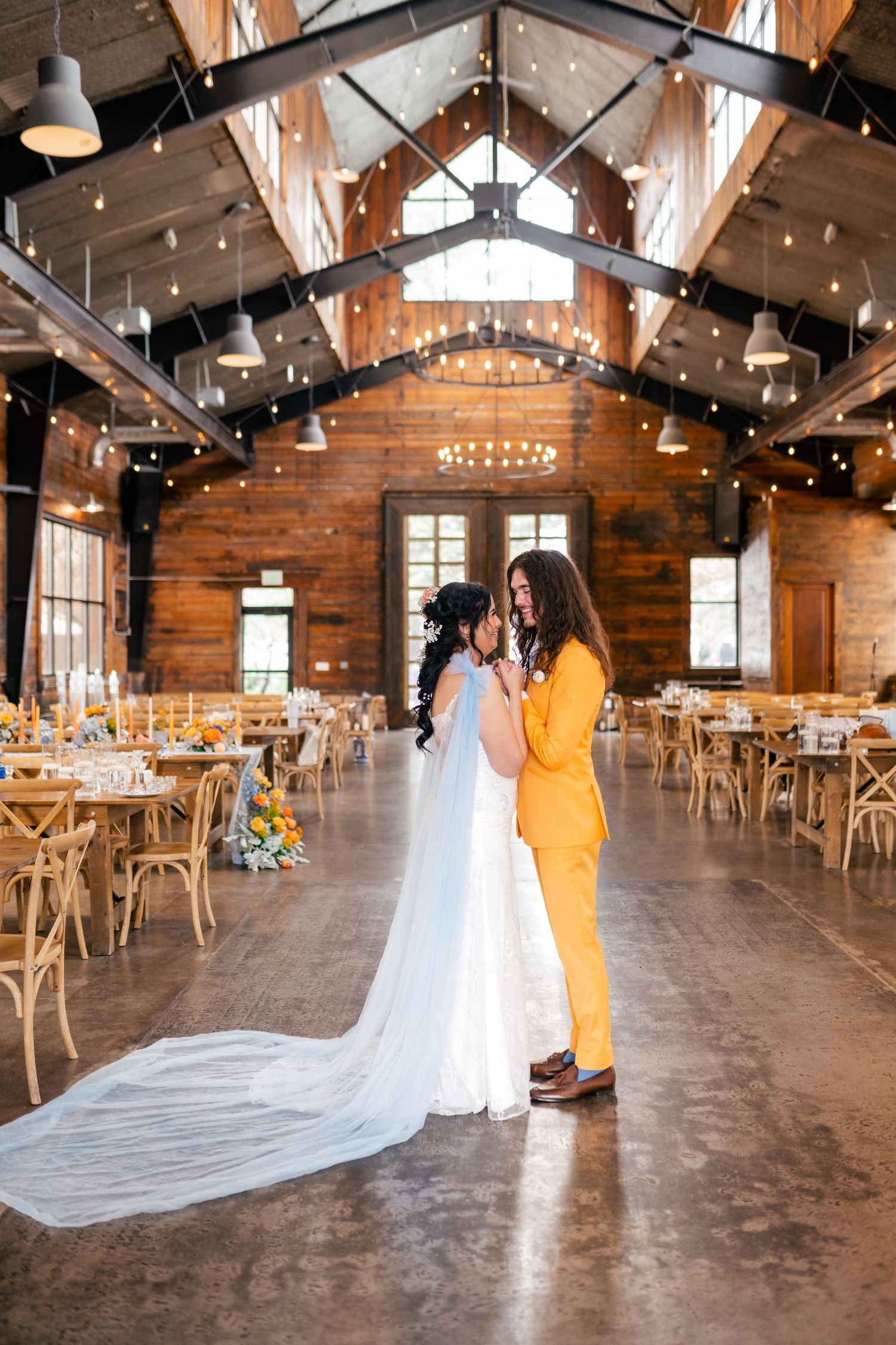 A flowing wedding veil dramatically trails across a rustic wooden venue floor with exposed beams and string lights overhead. Knot & Pine.