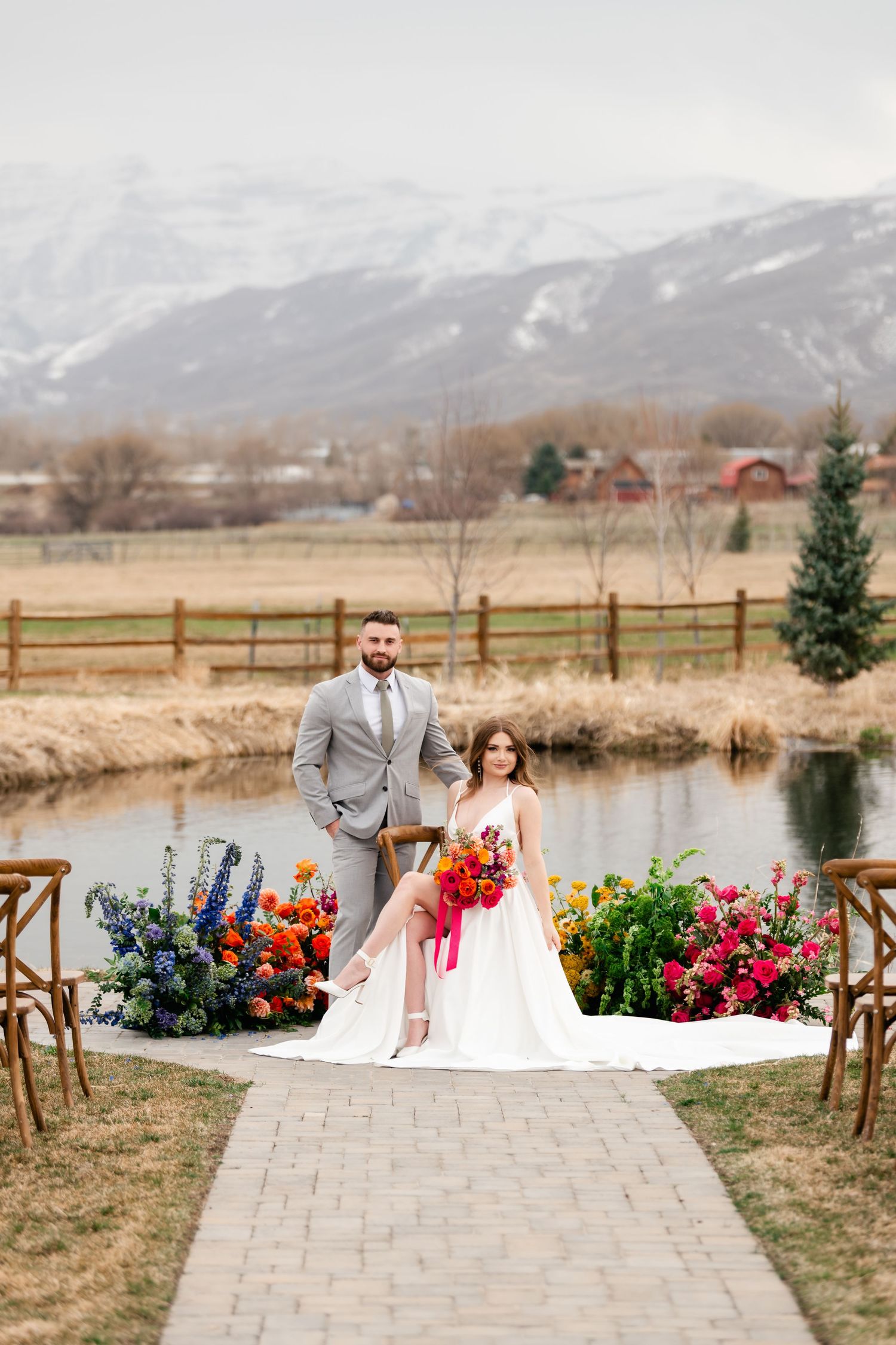A scenic outdoor wedding ceremony setup with colorful floral arrangements near a pond with mountains in background at River Bottoms Ranch.