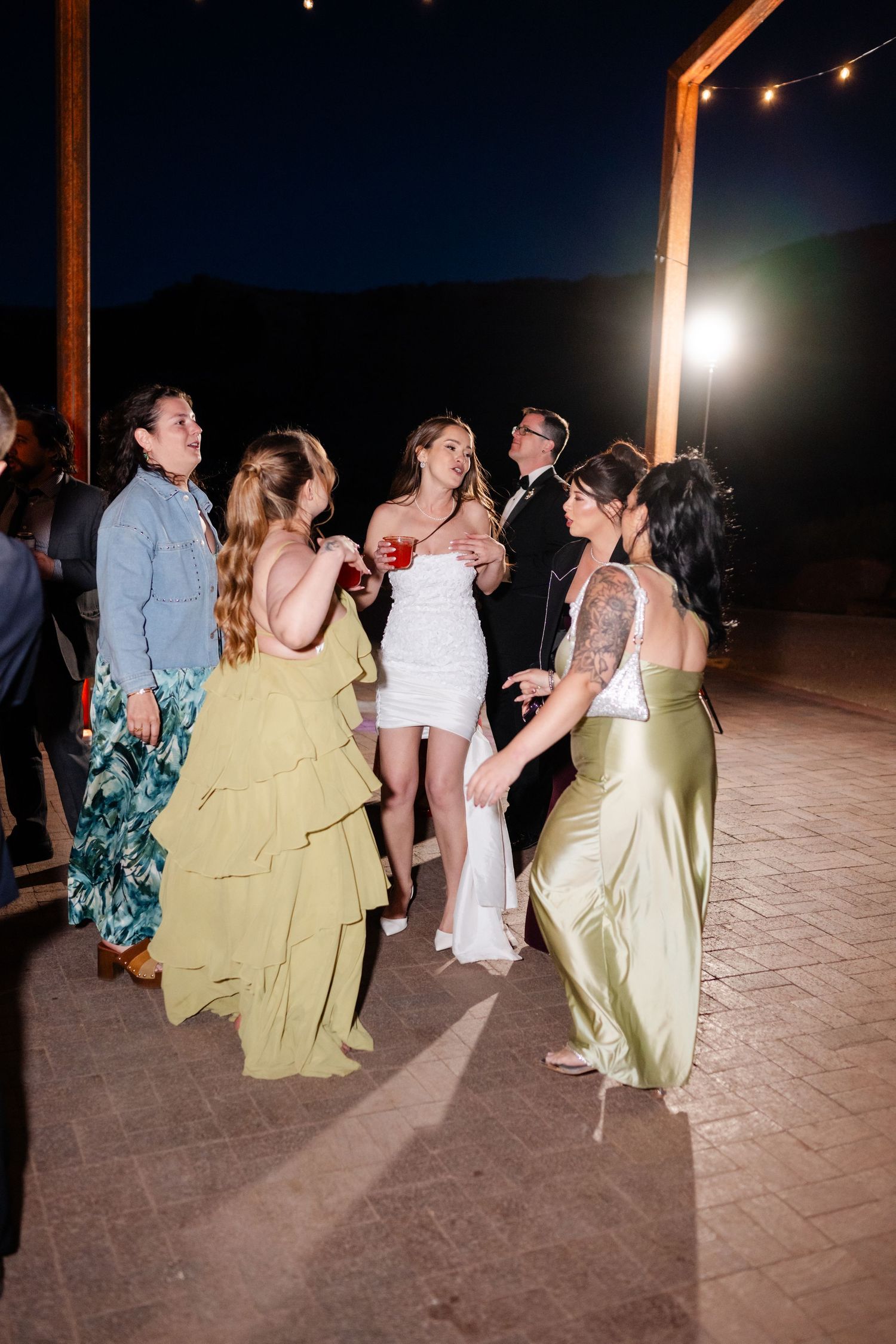 Women in formal gowns dancing outdoors at night wedding reception under string lights.