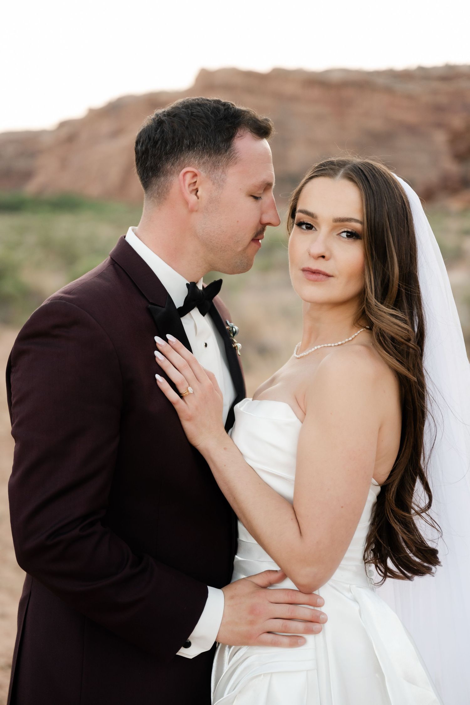 Groom in burgundy tuxedo tenderly kisses bride in strapless white gown with veil in desert setting.