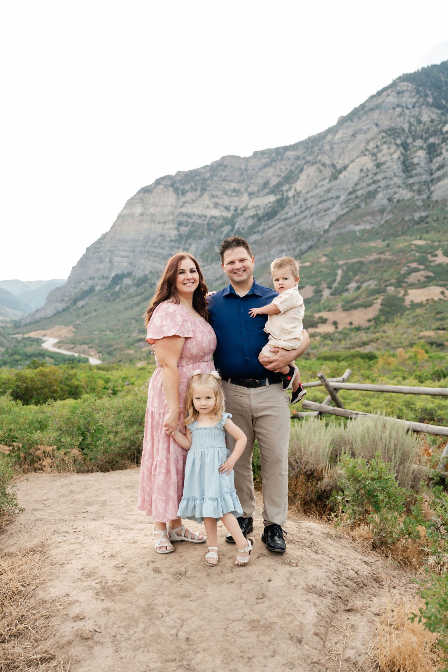 Family stands together on a scenic mountain trail with rugged cliffs and desert vegetation in the background.