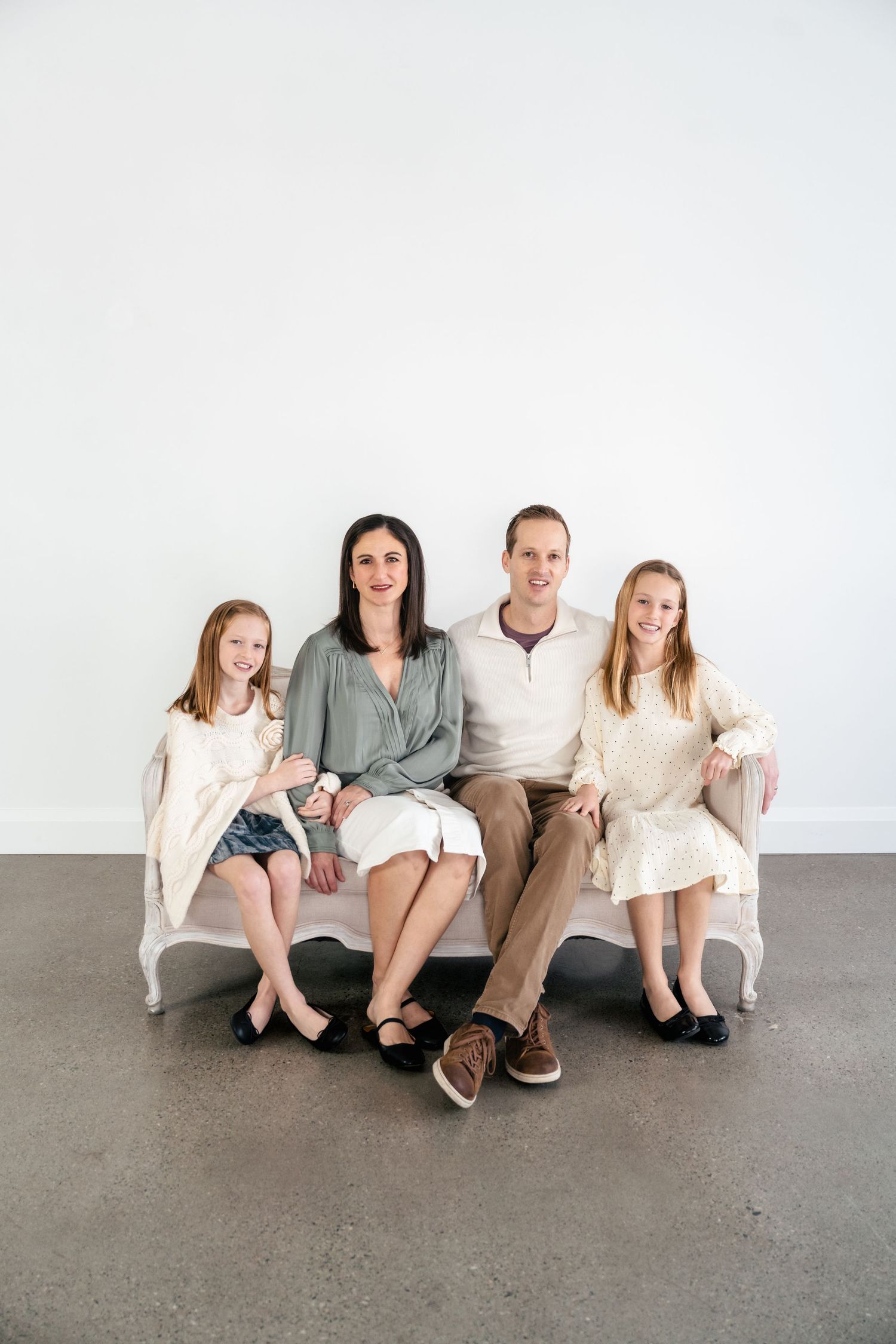 A family of four sits together on a modern white curved bench against a plain white wall.