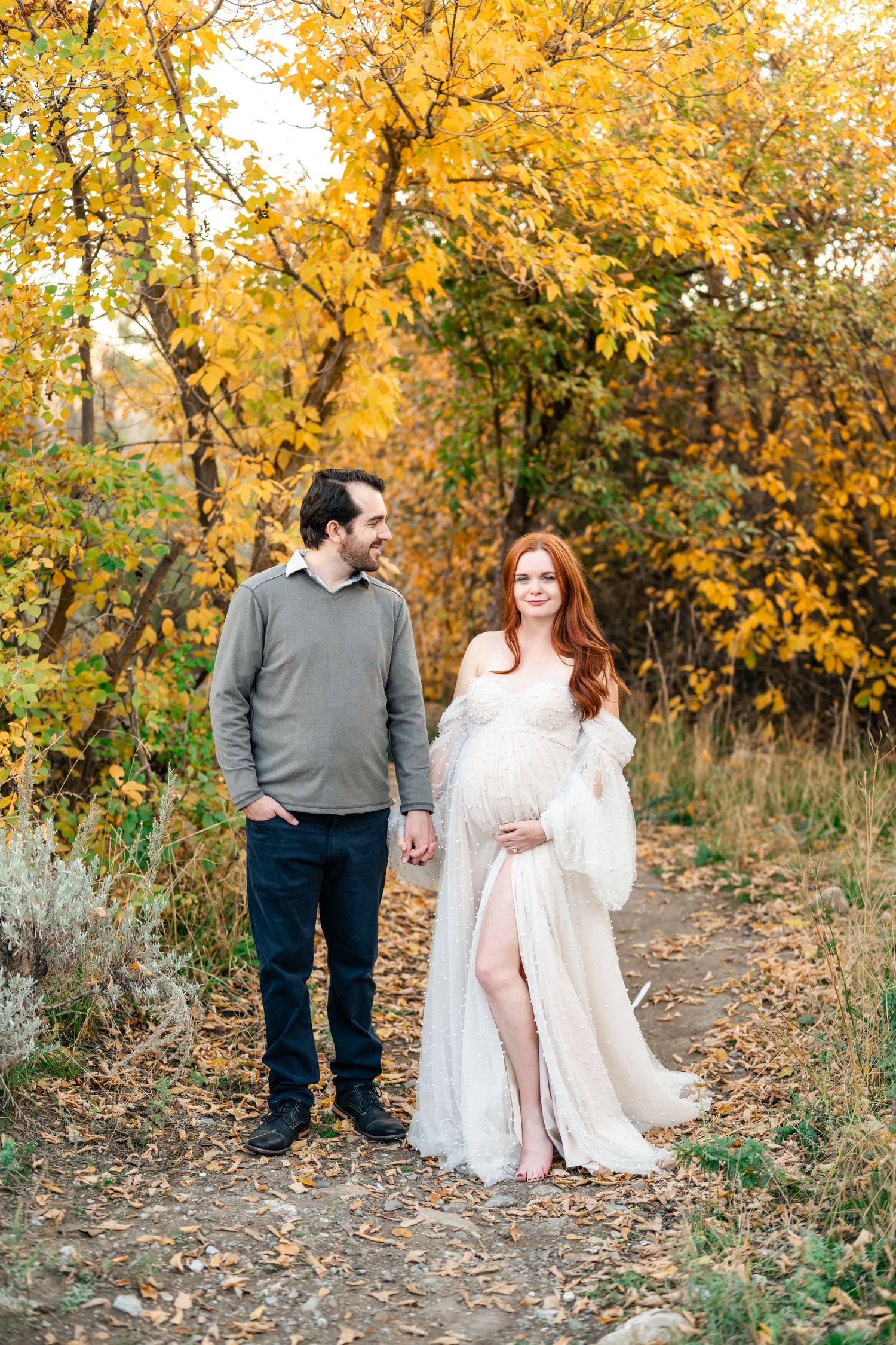 A couple stands together on an autumn path surrounded by golden yellow trees and fallen leaves.