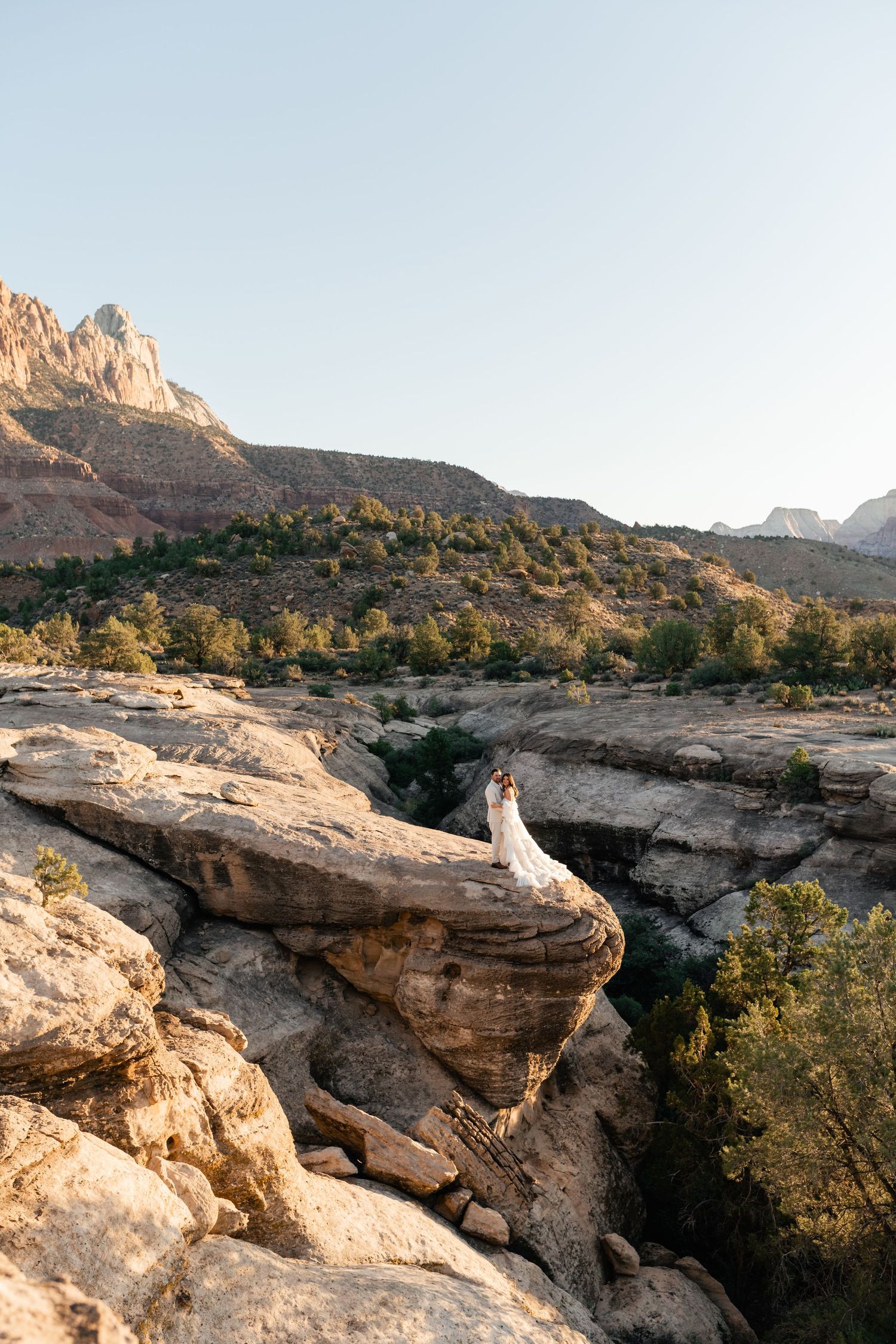 Zion Adventure Session. Wedding photos captured on a dramatic rocky cliff overlook with desert mountains and blue sky in the distance.