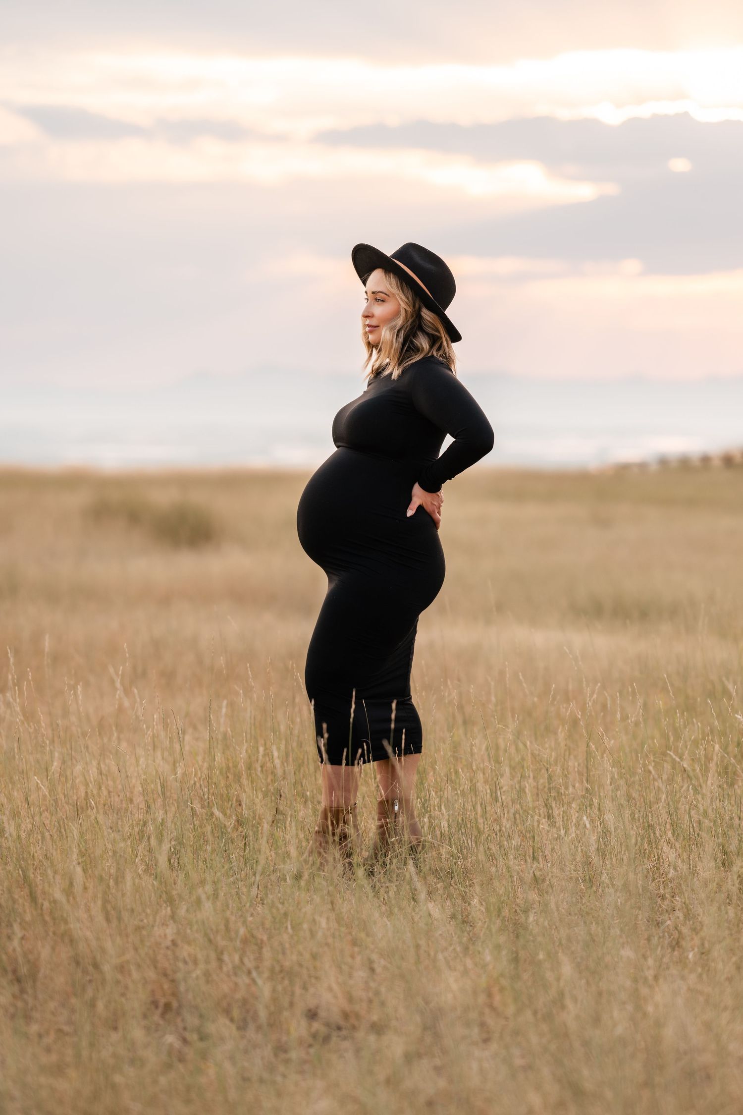 Artistic maternity portrait in black dress and hat against golden field at sunset.