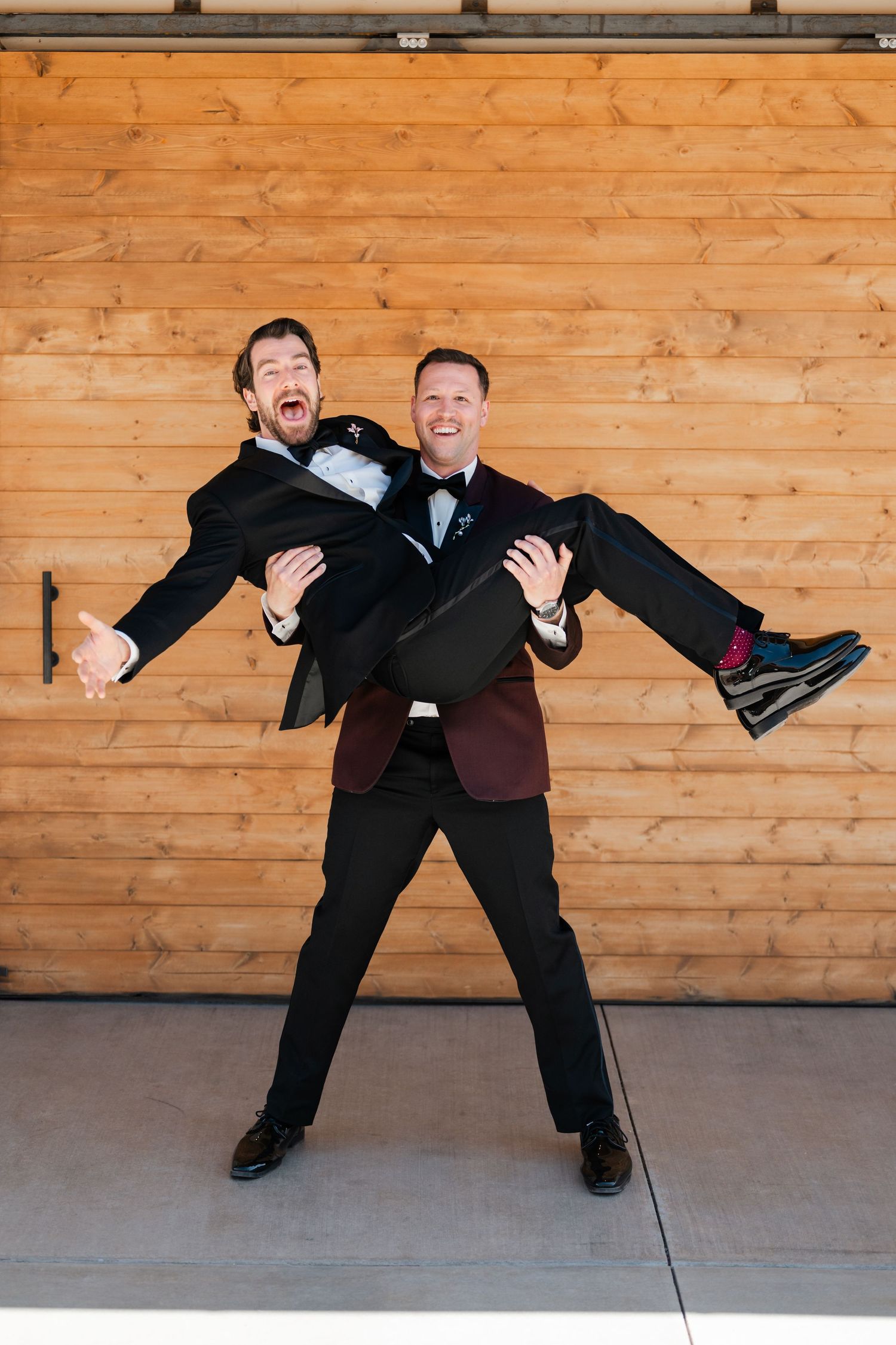 Two grooms laugh joyfully as one carries the other in his arms outside a wooden barn at their wedding.