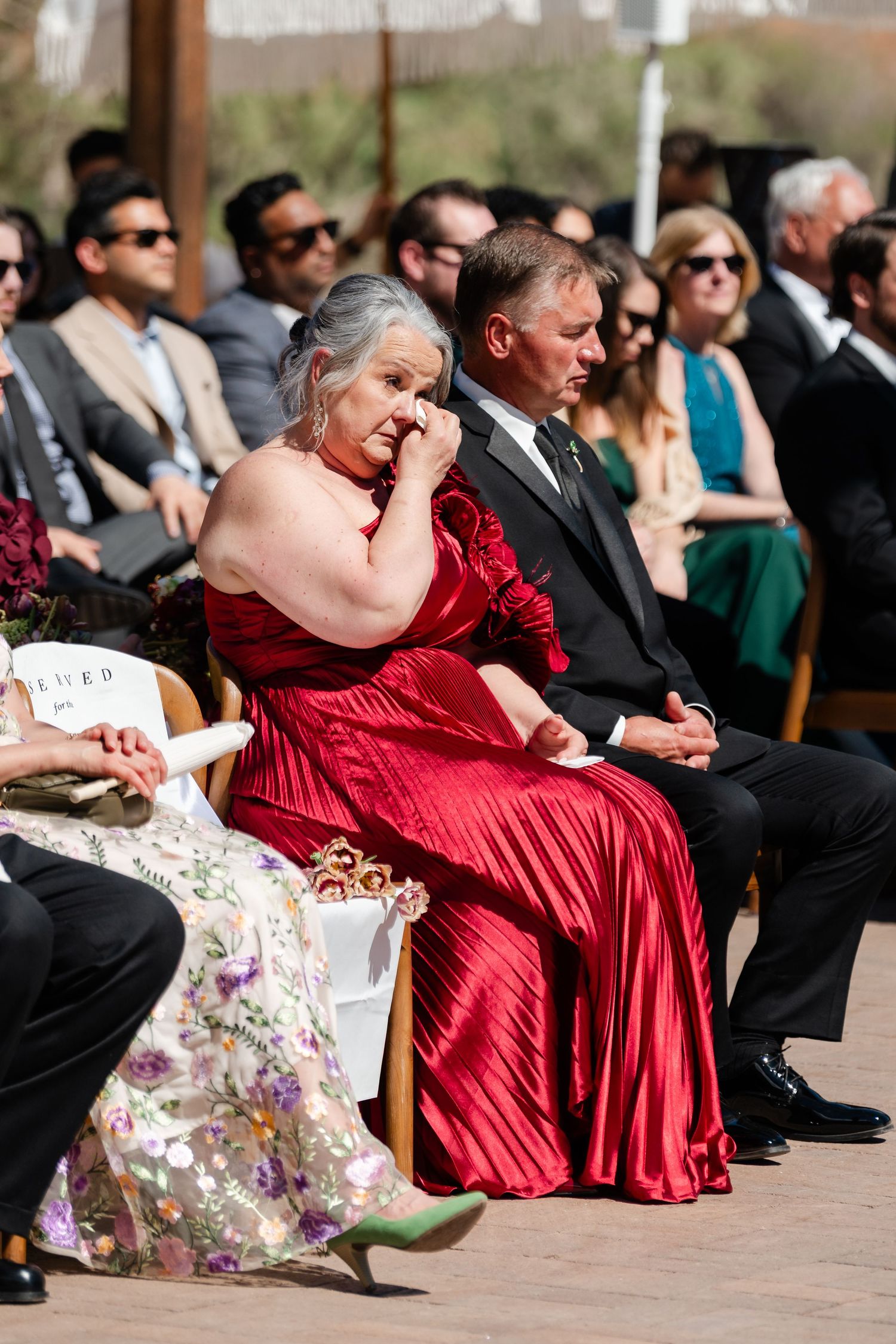 Emotional wedding guest in red satin gown wiping tears during outdoor ceremony, seated among formally dressed attendees.