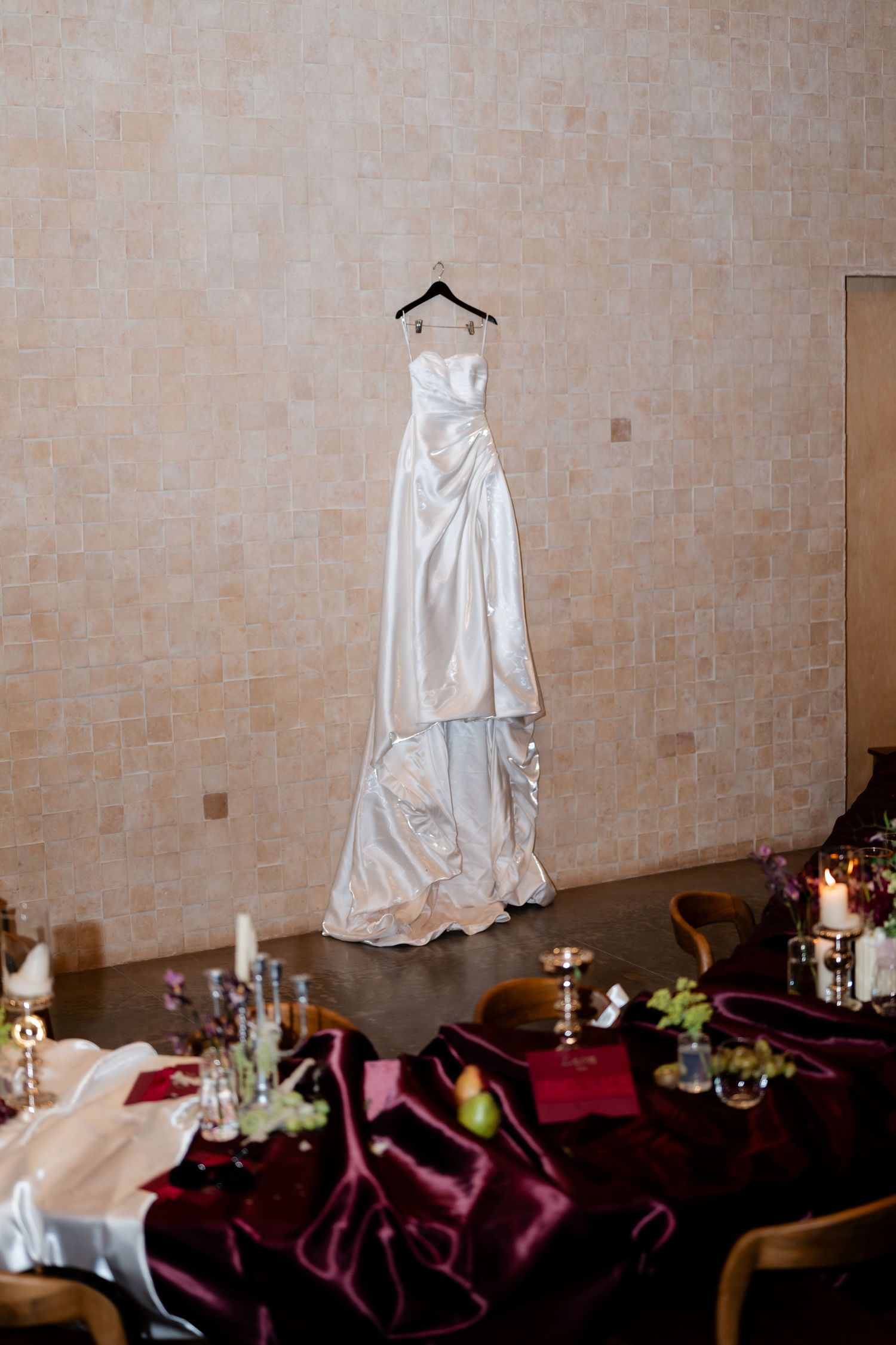 Elegant white satin wedding gown hanging on a hanger against a tiled wall above a decorated reception table.
