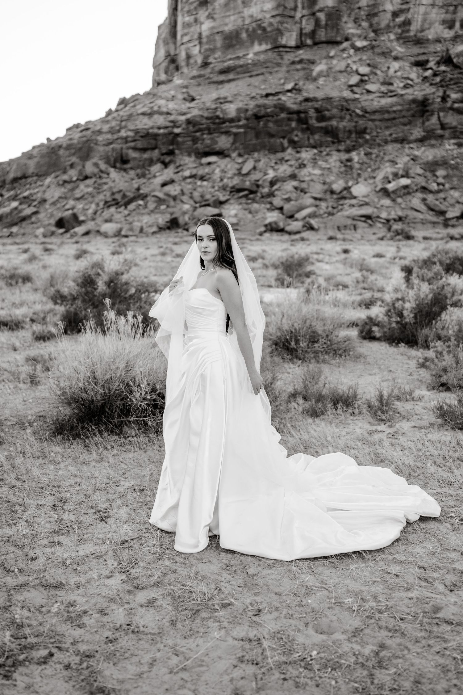 Black and white photo of a bride in a strapless gown with veil in a desert landscape with red rock formations.