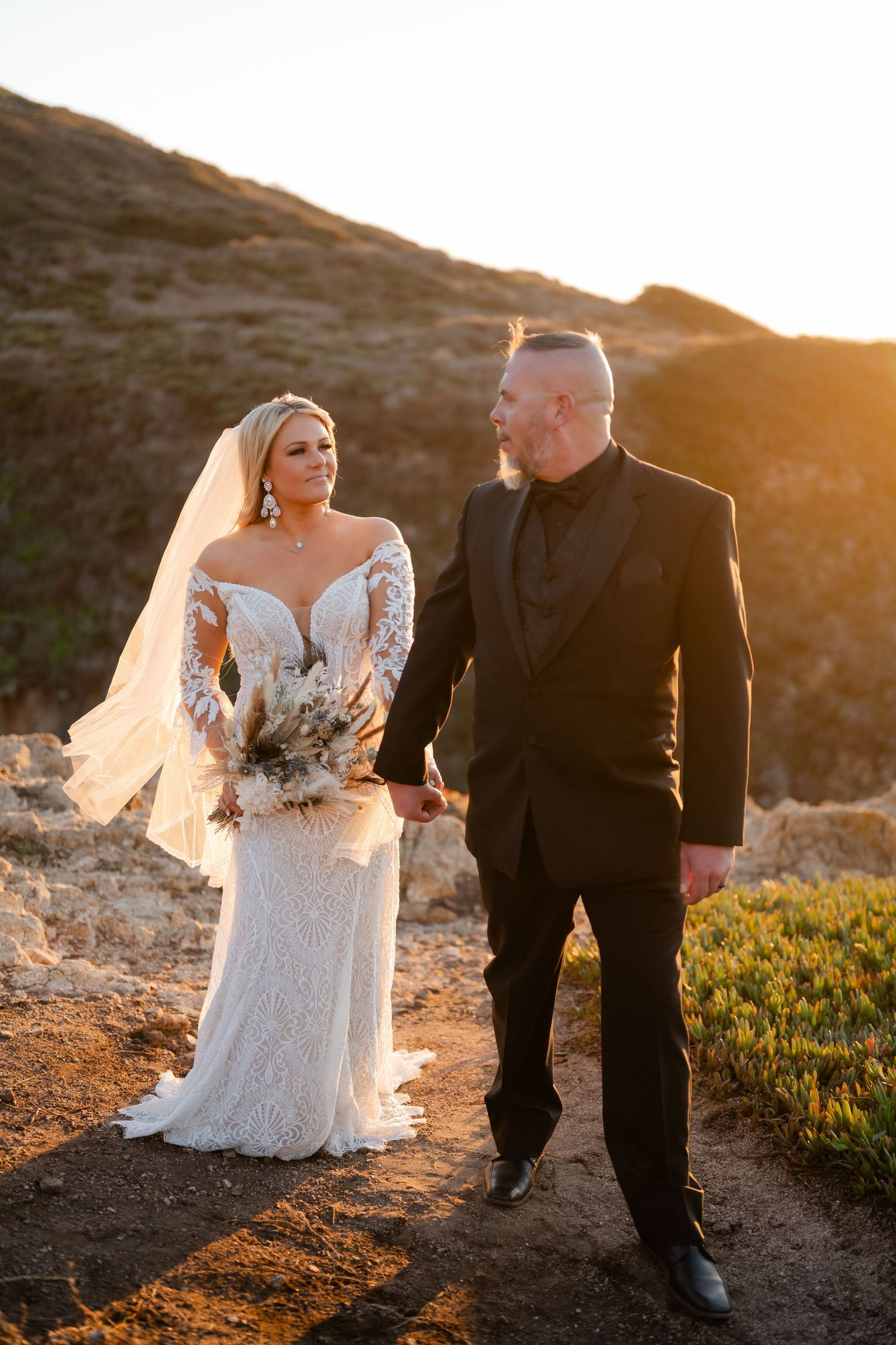Bride and groom walking together at sunset on a hillside with golden light illuminating their silhouettes.