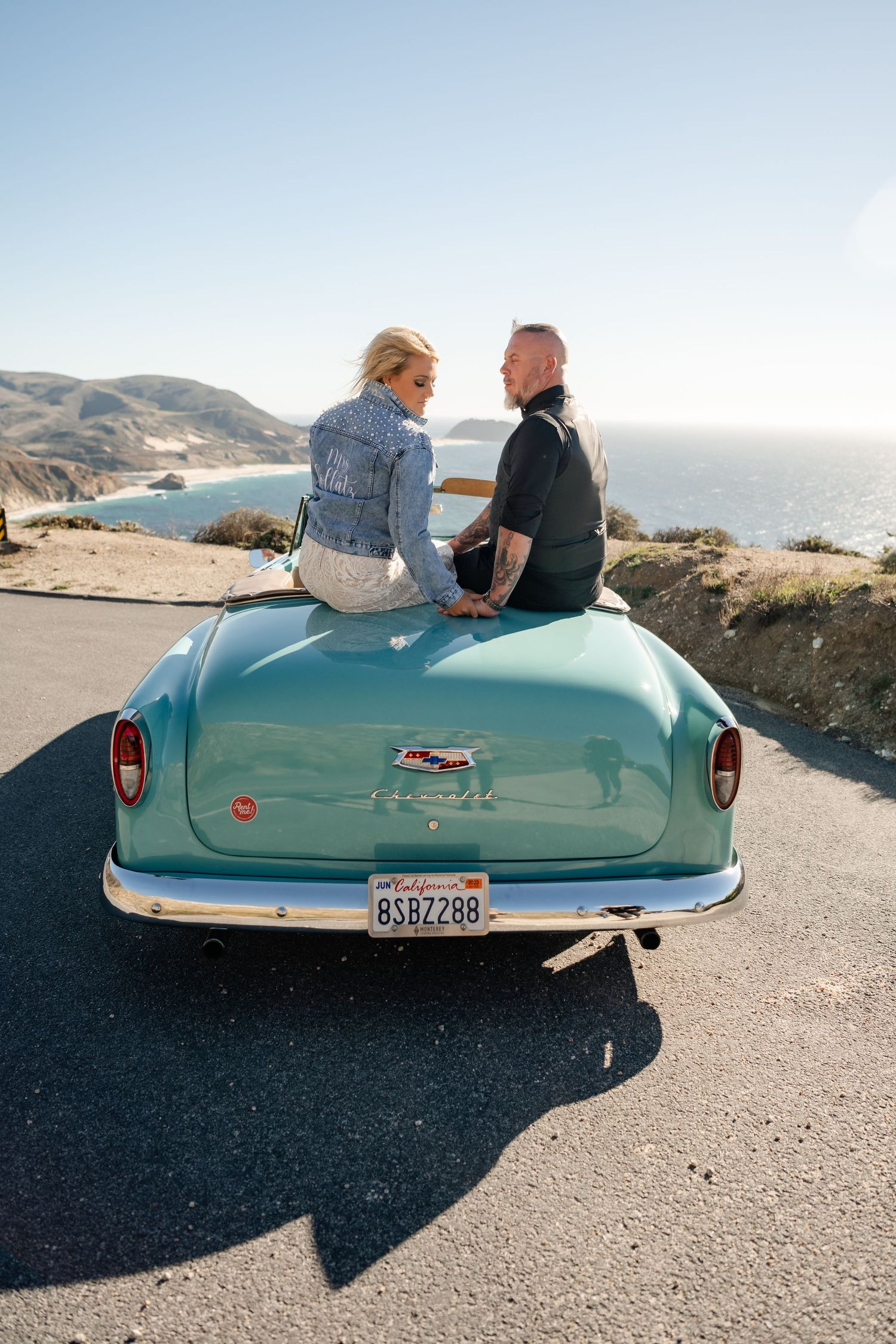 A couple sits on a vintage turquoise car overlooking the ocean on a sunny day.