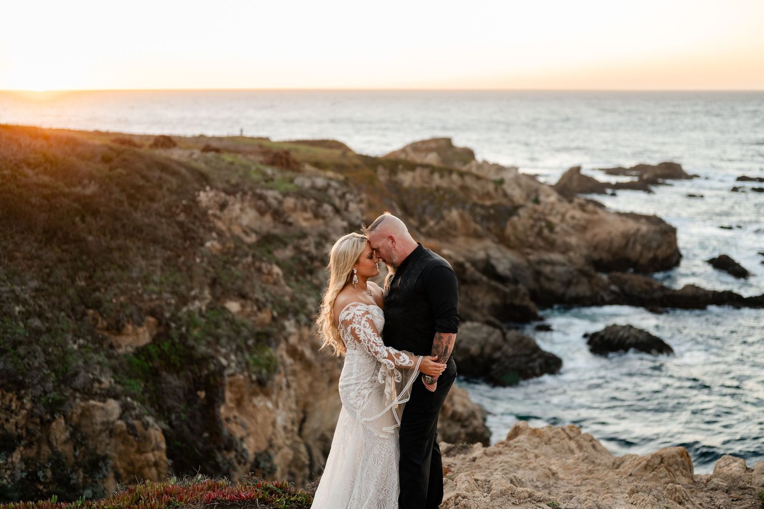 A romantic wedding photo on rocky coastal cliffs during golden hour.