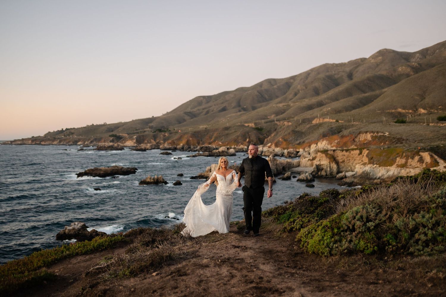 A wedding ceremony takes place on a scenic cliff overlooking the rugged Big Sur coastline at sunset.