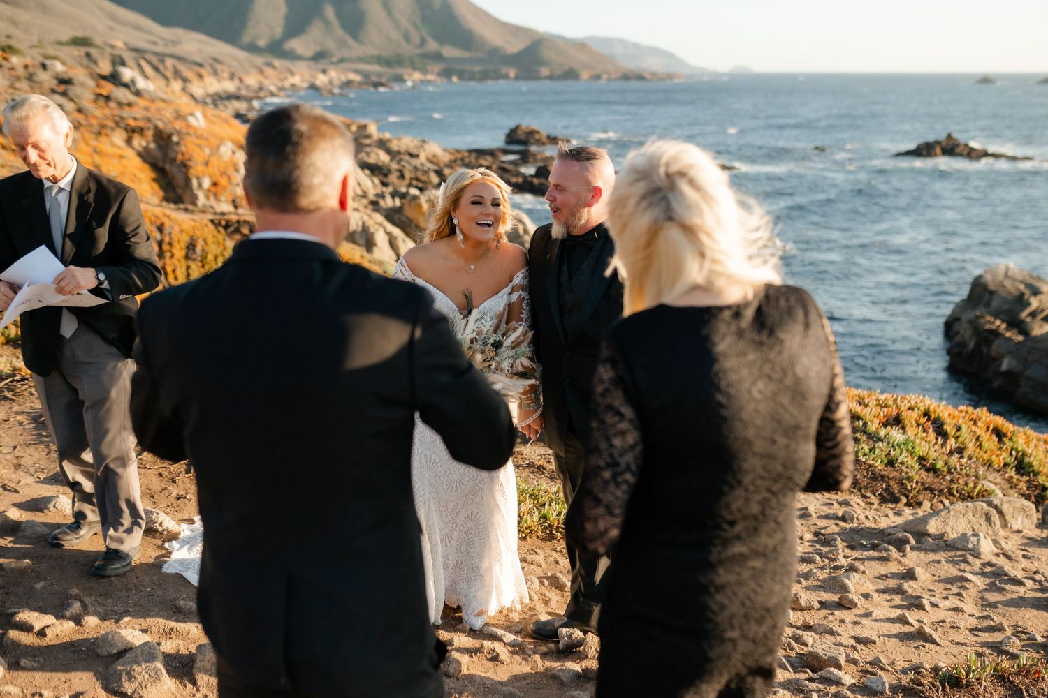 Wedding guests share joyful moments during an intimate ceremony on a coastal cliff.