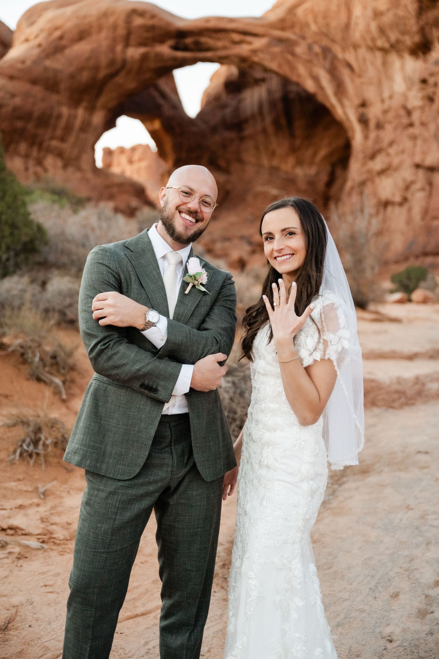 A happy wedding couple poses beneath a natural stone arch in a desert landscape.