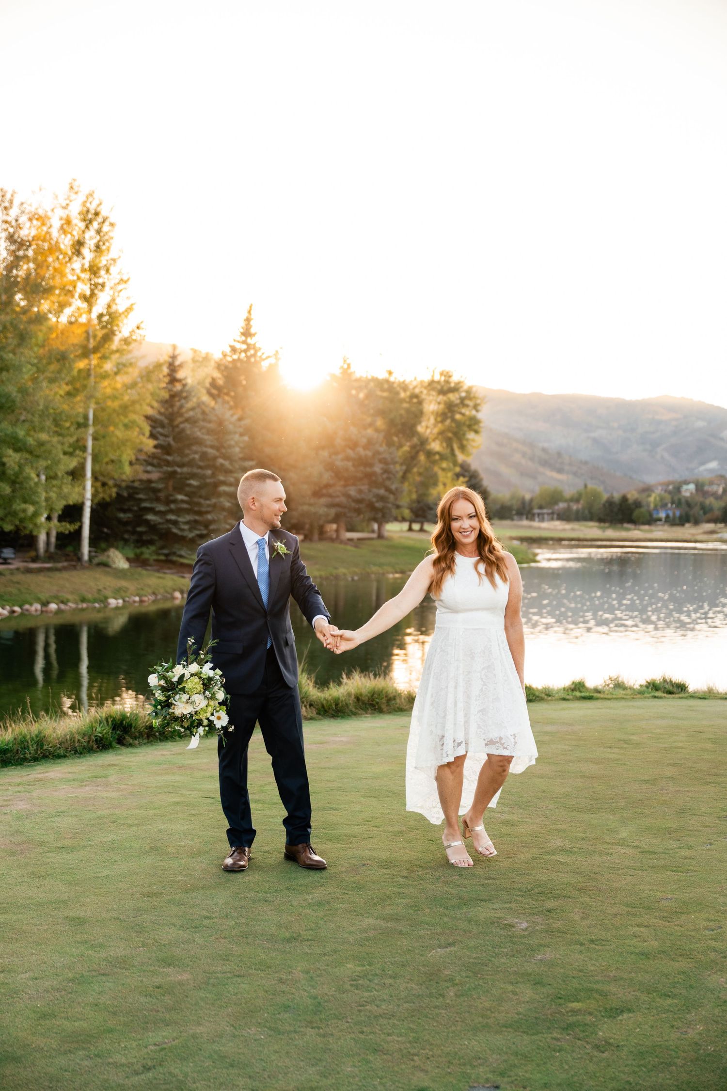 A couple shares a romantic moment on a lakeside golf course at sunset with mountains in the background.