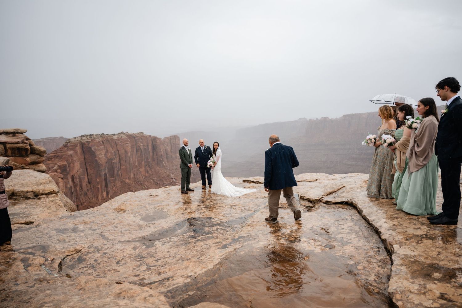Wedding guests watch ceremony unfold on dramatic cliff edge in overcast weather.