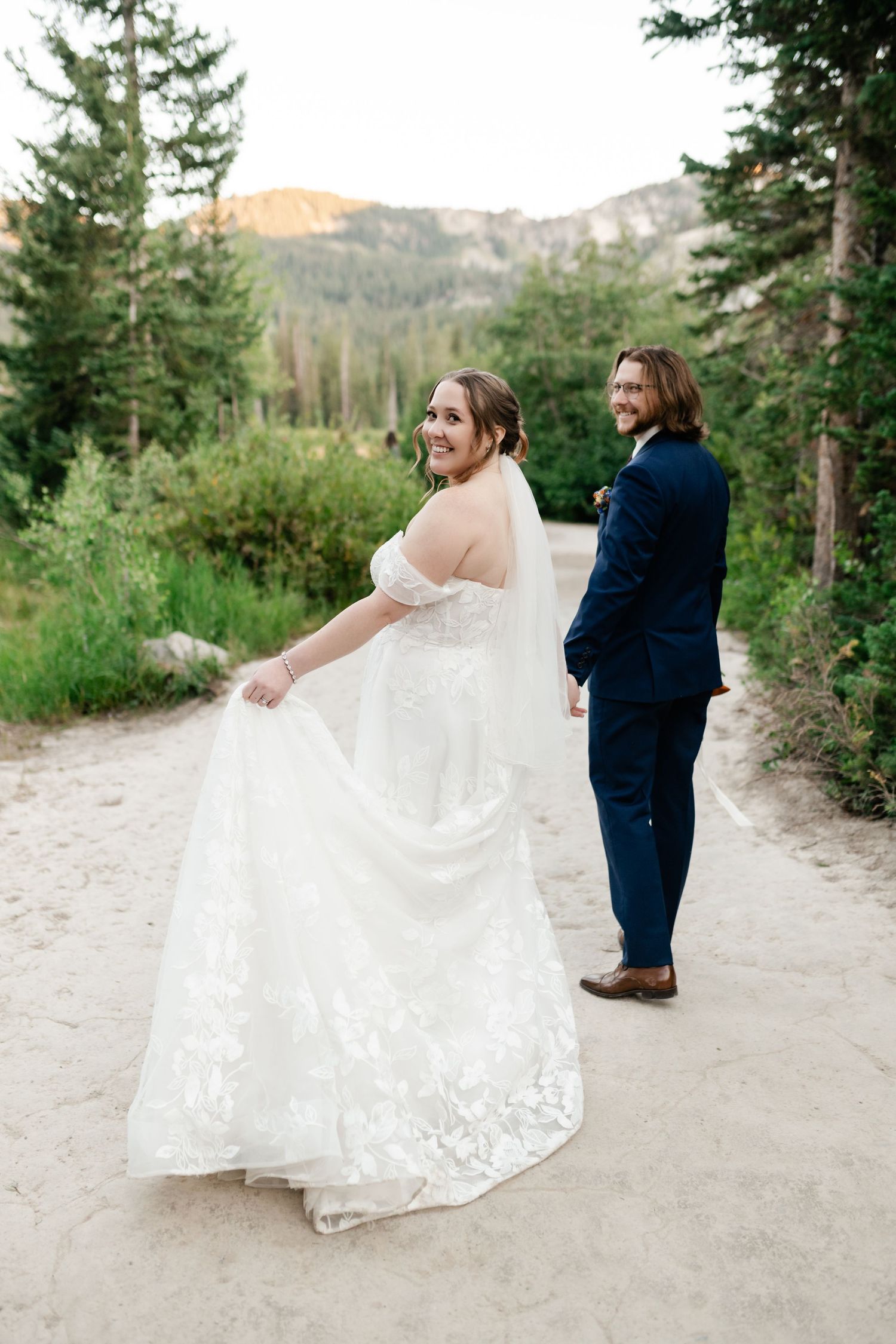 A couple in wedding attire dances playfully on a mountain path surrounded by evergreen trees and scenic views. Utah Mountain Wedding.