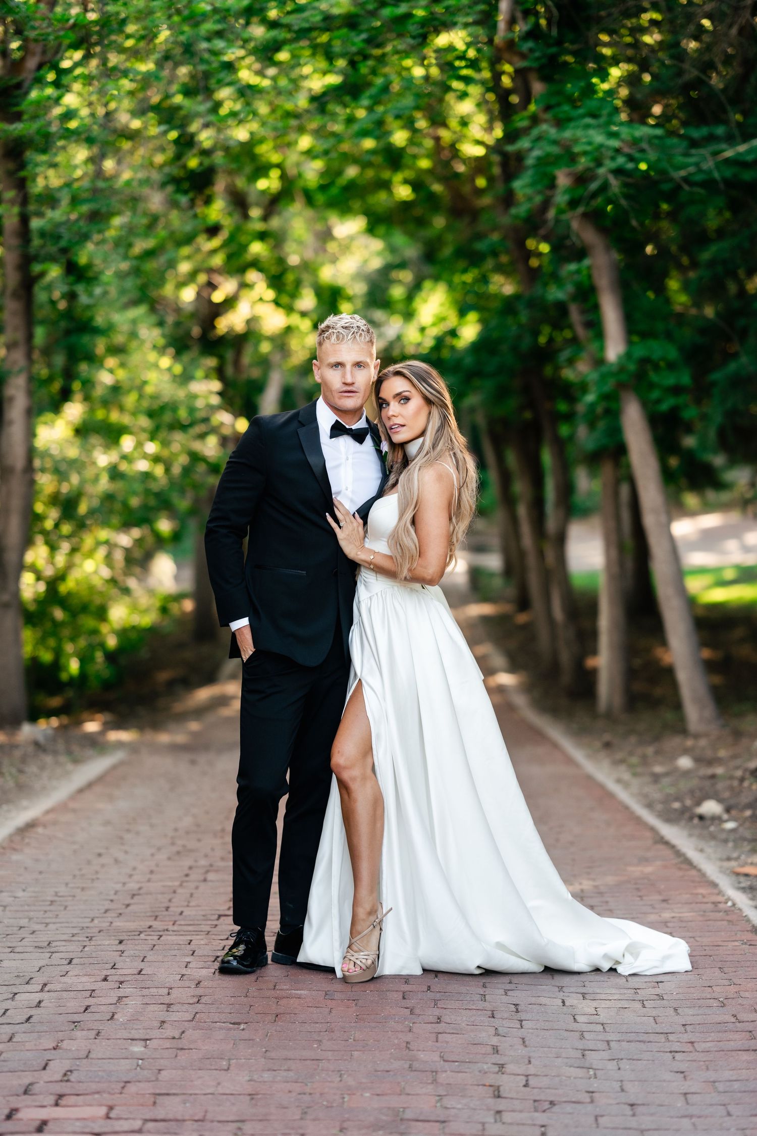 Wedding couple posing on brick pathway surrounded by lush green trees.