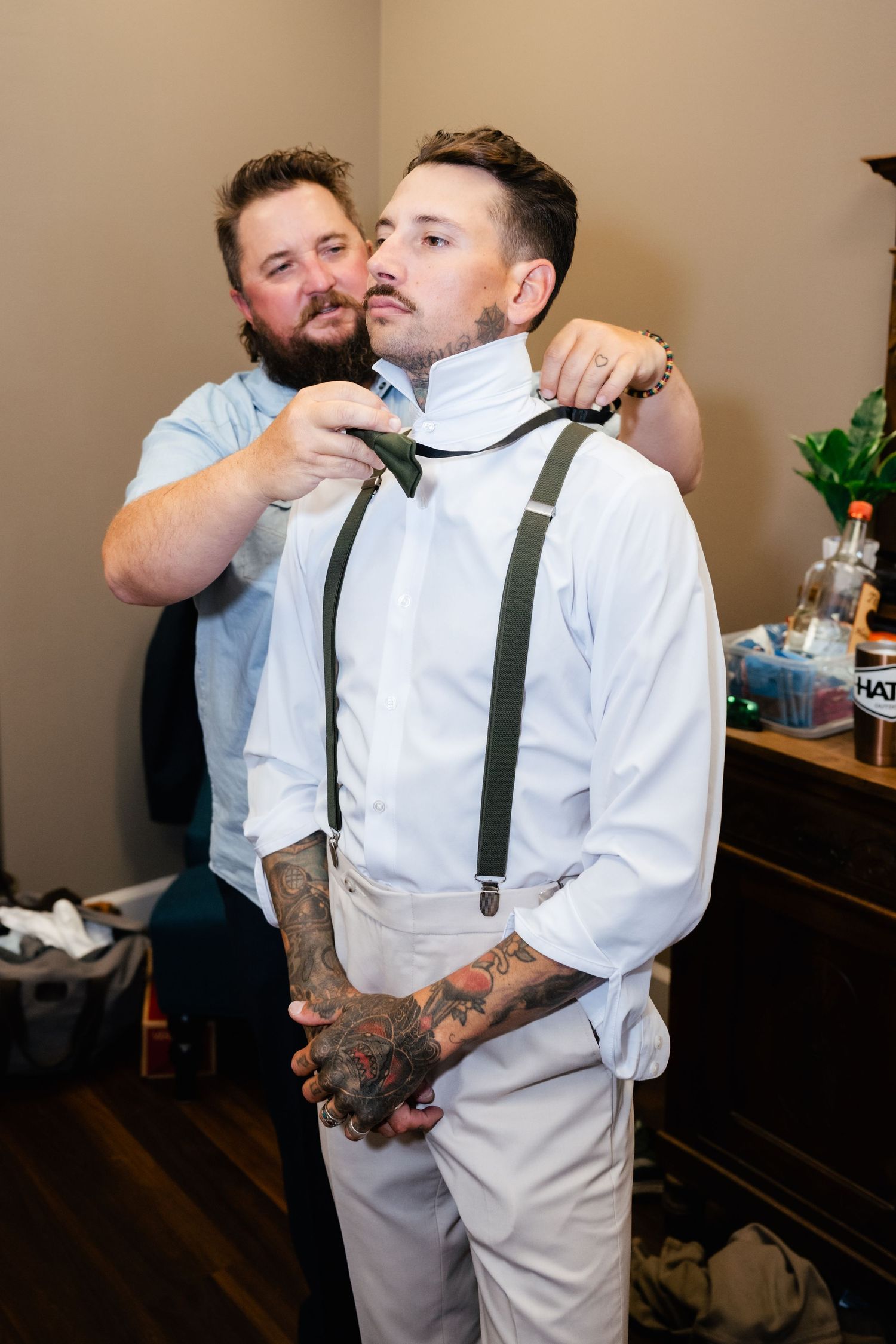 Two people prepare for a wedding ceremony as suspenders are adjusted in a getting-ready room.