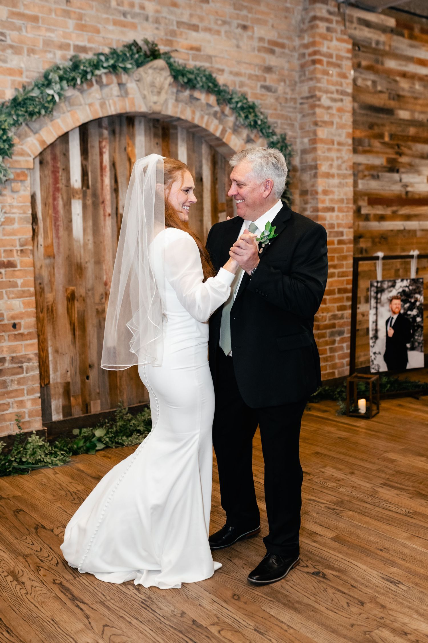 A couple shares a sweet dance moment together at their wedding venue with a brick wall and greenery arch backdrop.