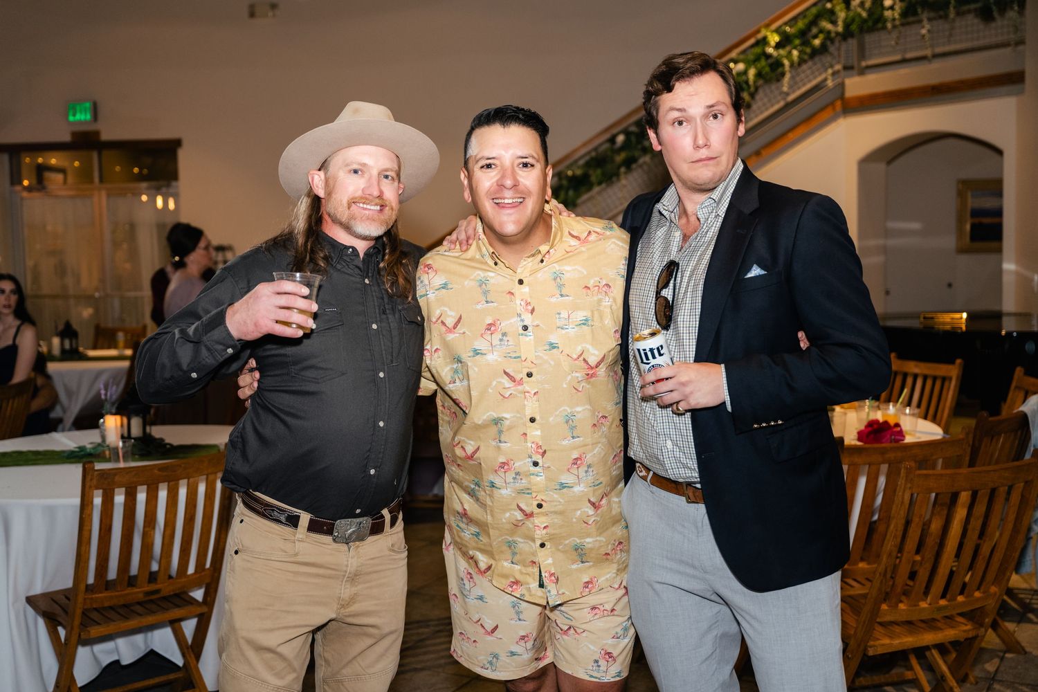 Three guests pose with drinks at a wedding reception in casual and formal attire.