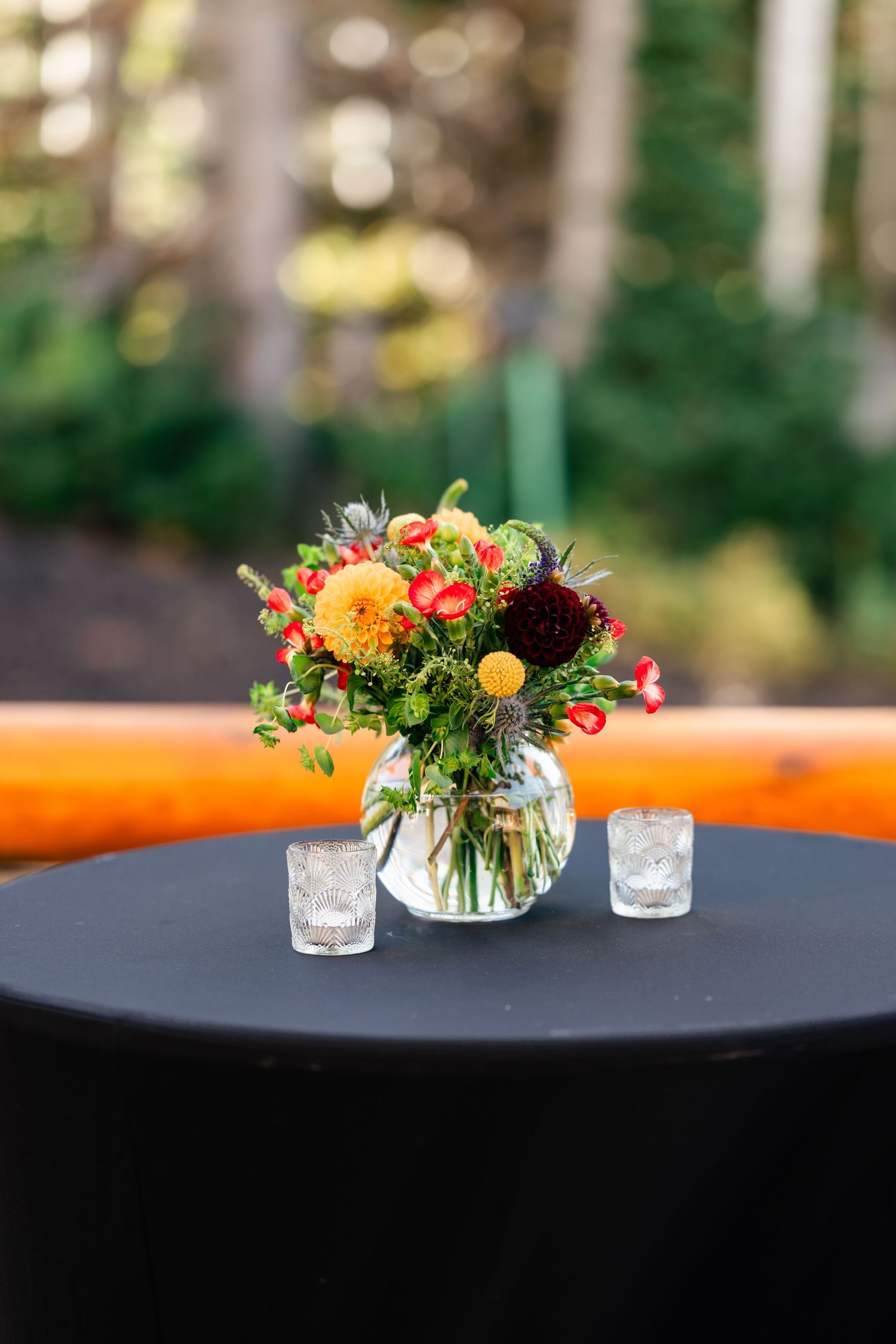 Colorful floral centerpiece with orange and dark red flowers in glass vase on black tablecloth.