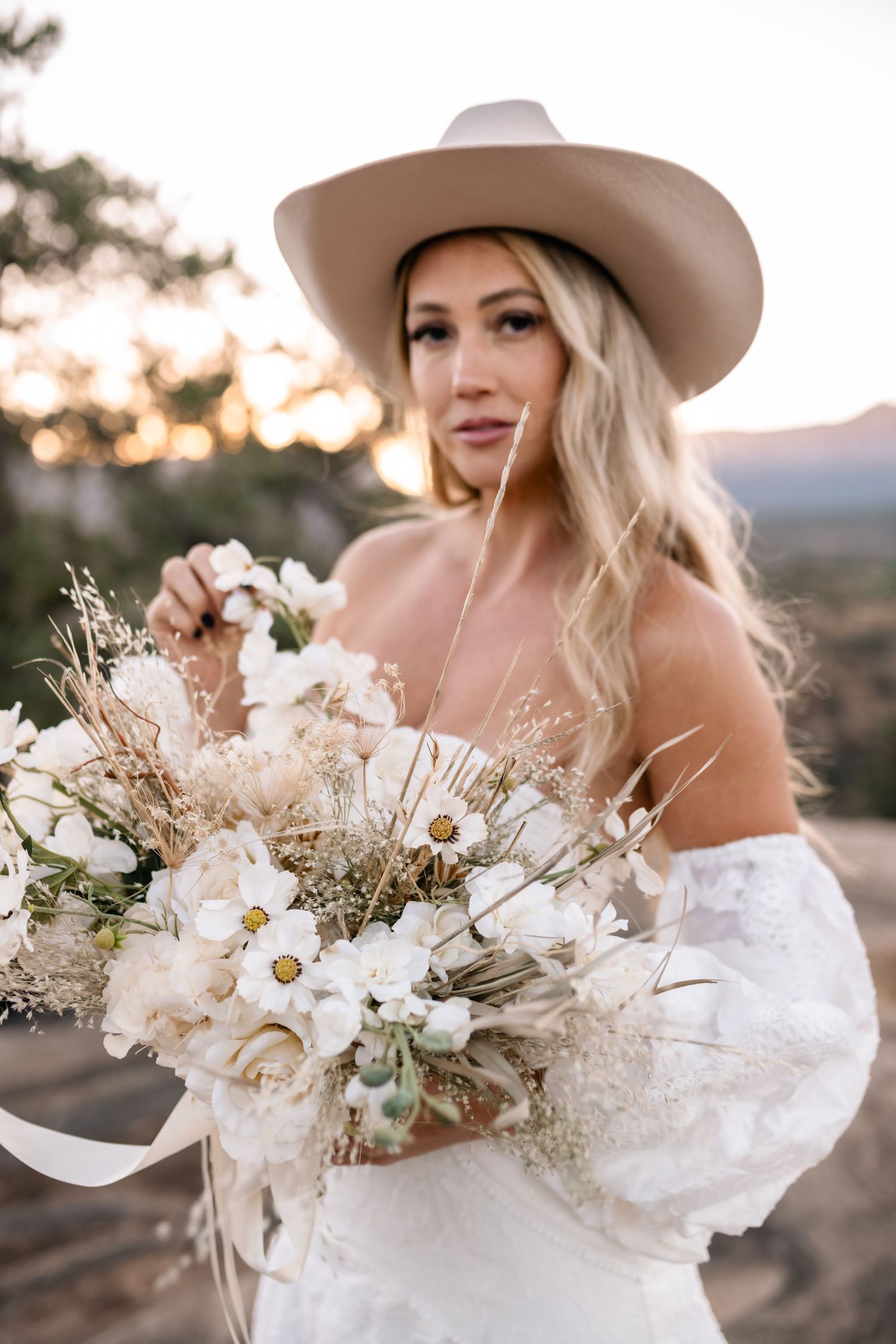 Dried floral wedding bouquet with neutral tones held against mountain sunset backdrop.