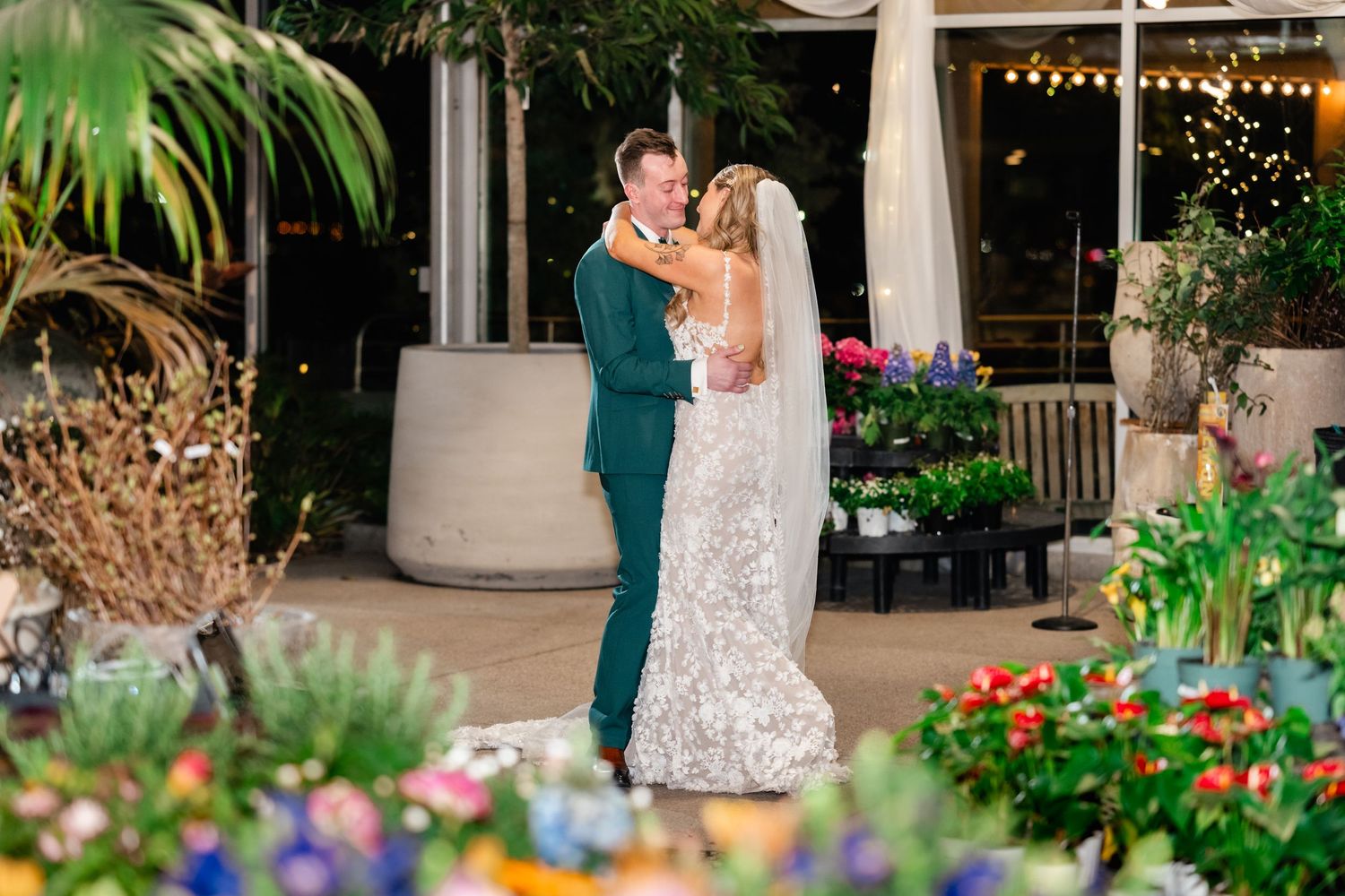 A couple shares a romantic moment on their wedding day surrounded by colorful flowers and twinkling lights at a tropical venue.