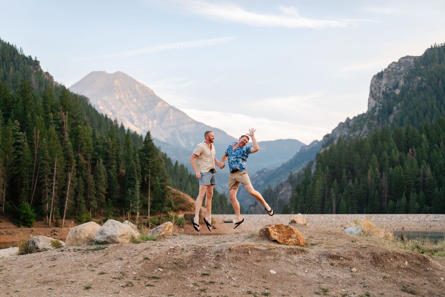 Two friends jumping joyfully outdoors with mountain and pine forest backdrop on a clear day.