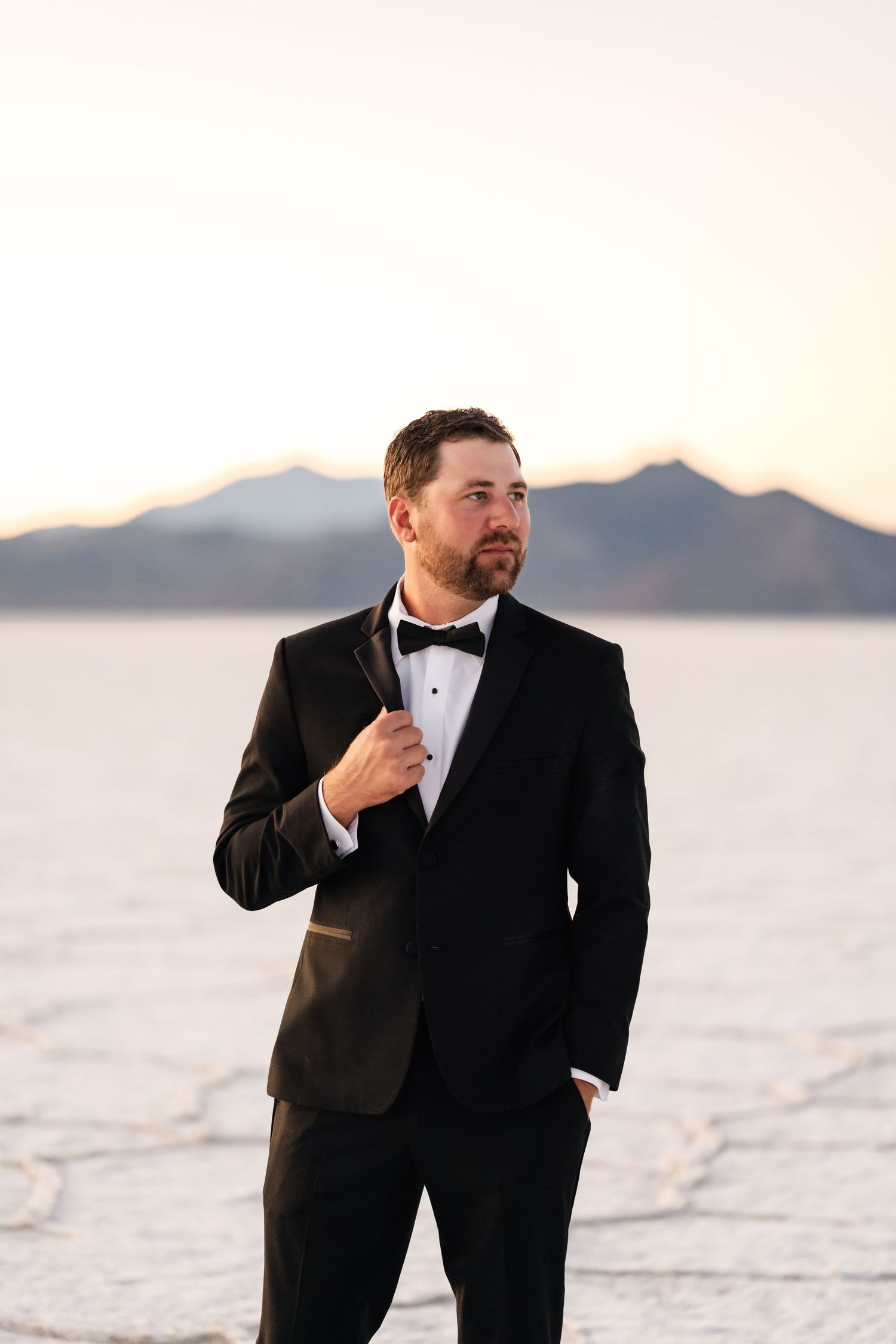 Utah Salt Flats Photographer. A person in a black tuxedo and bow tie poses against a scenic mountain and salt flat landscape at sunset.