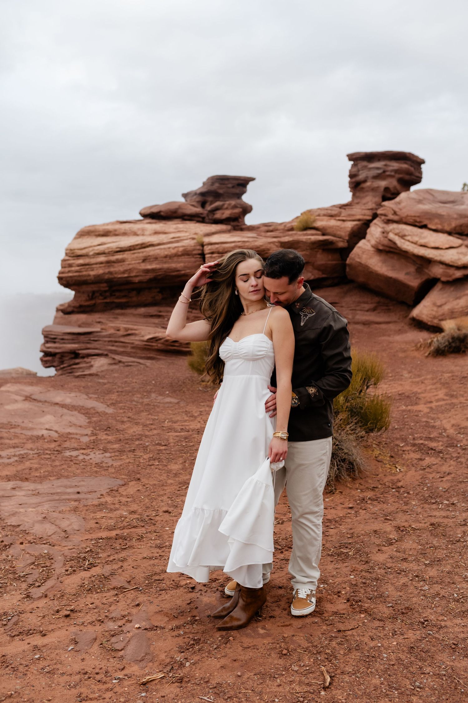 A romantic moment captured between a couple on red desert rocks under cloudy skies.