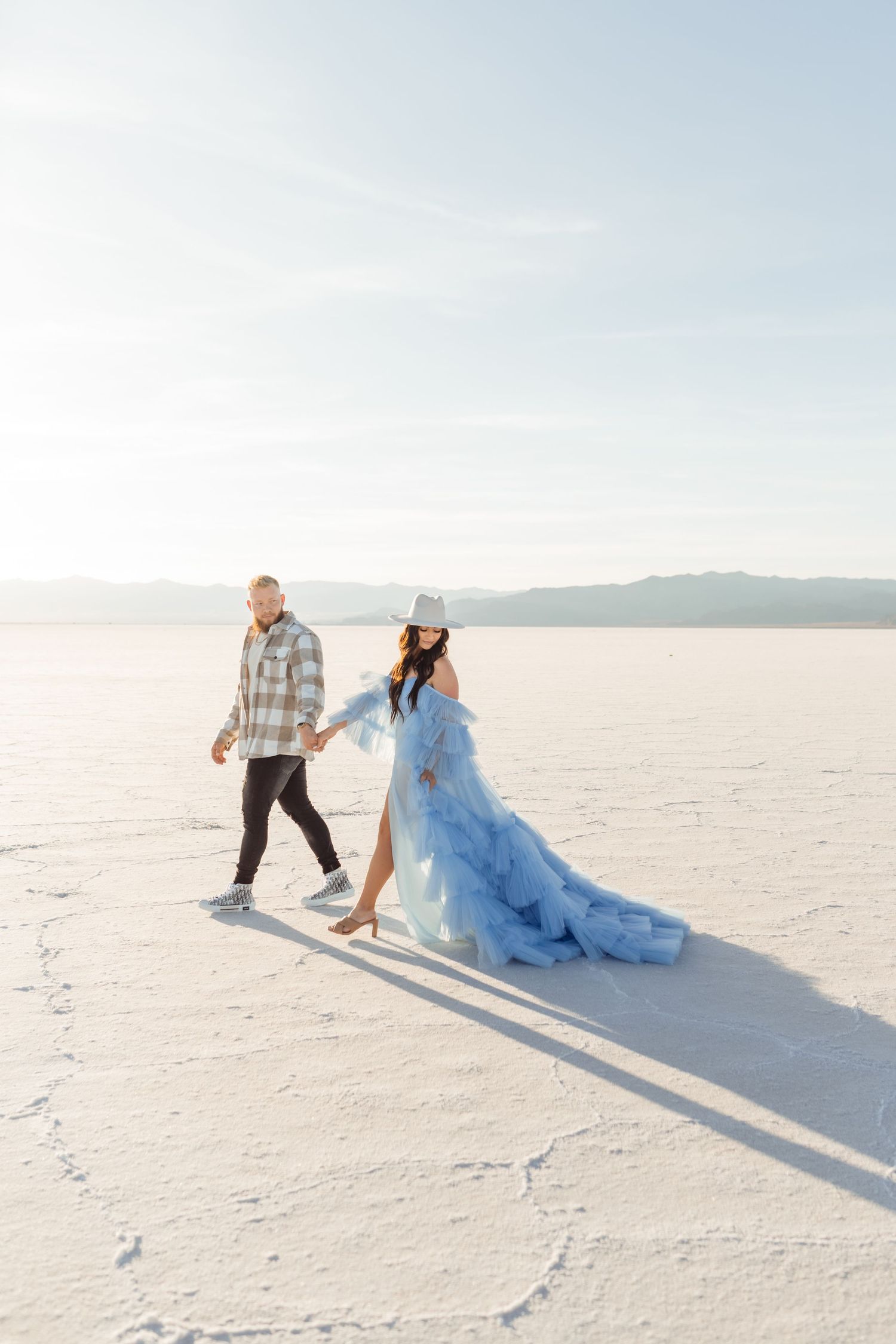 Utah Salt Flats Photos. A couple walks hand in hand across a stark white salt flat landscape as her flowing blue dress trails behind them.