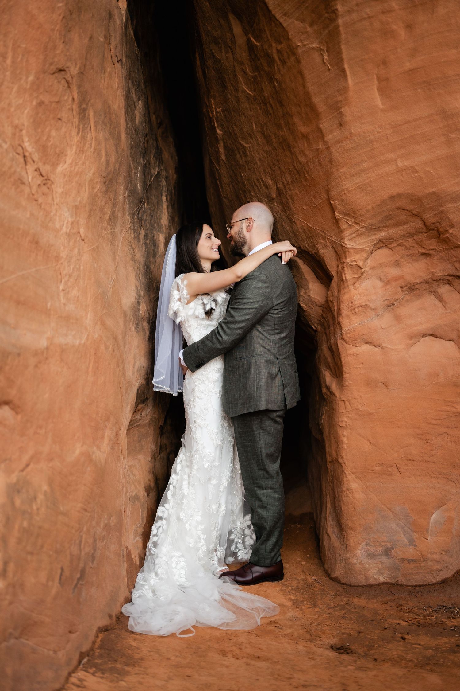 A couple embraces in a narrow sandstone canyon alcove wearing formal wedding attire.