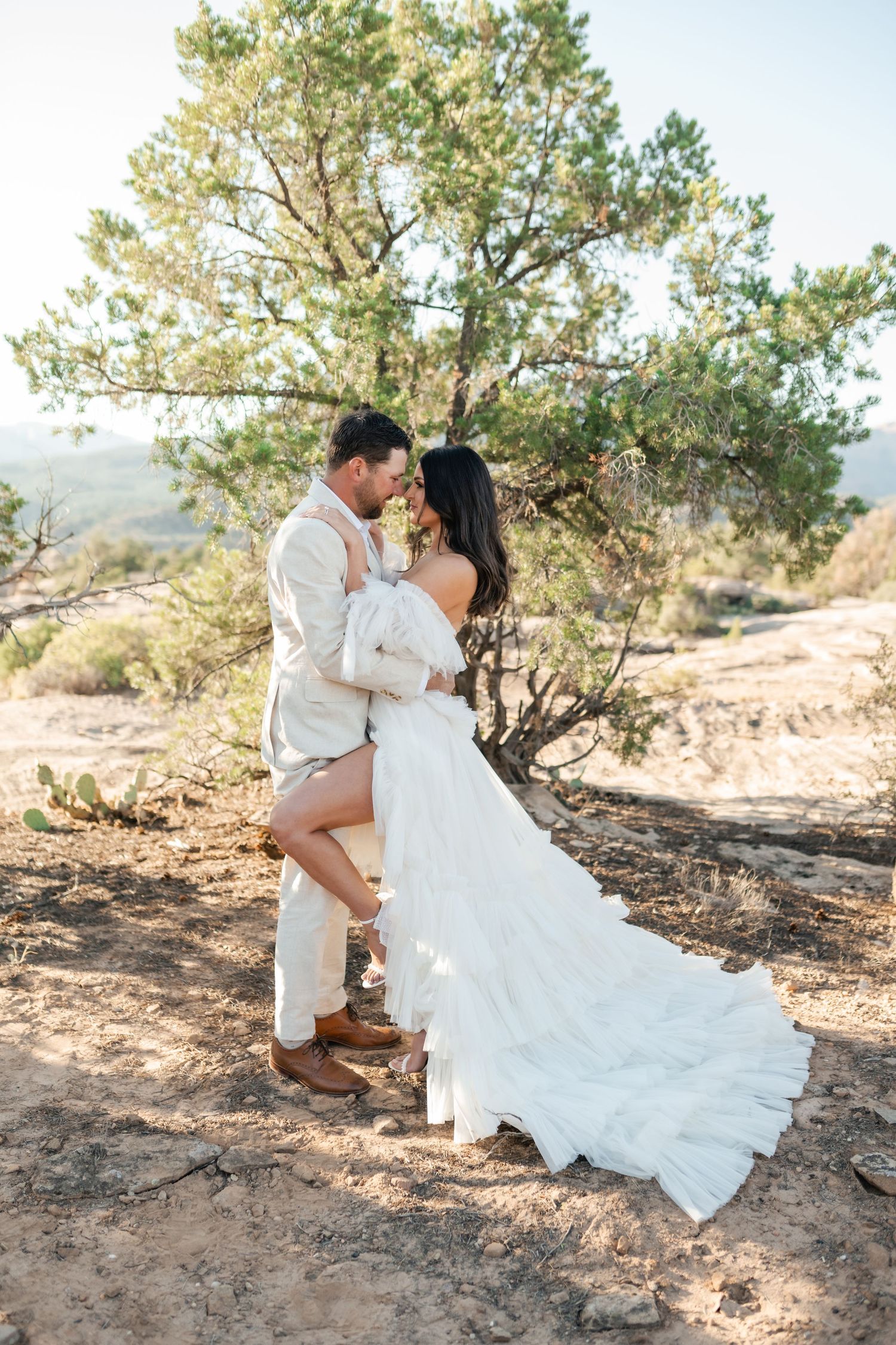 Zion destination photographer, A romantic couple sits together on desert terrain surrounded by pine trees in an intimate outdoor setting.