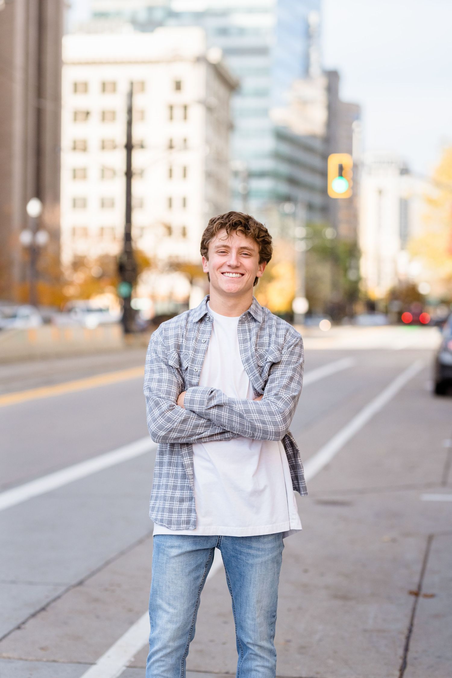 Young person in plaid shirt and jeans poses confidently on city sidewalk with urban buildings in background.