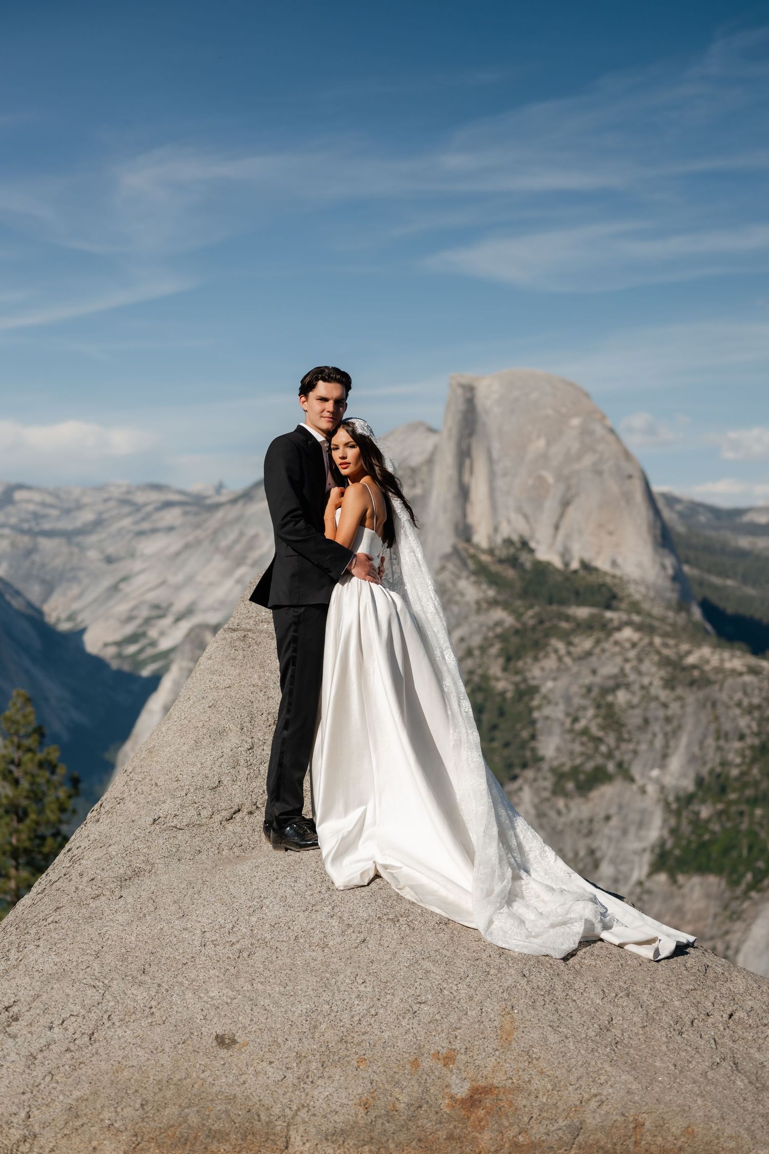 Wedding couple embraces on granite rock at Glacier Point with Half Dome mountain backdrop in Yosemite.