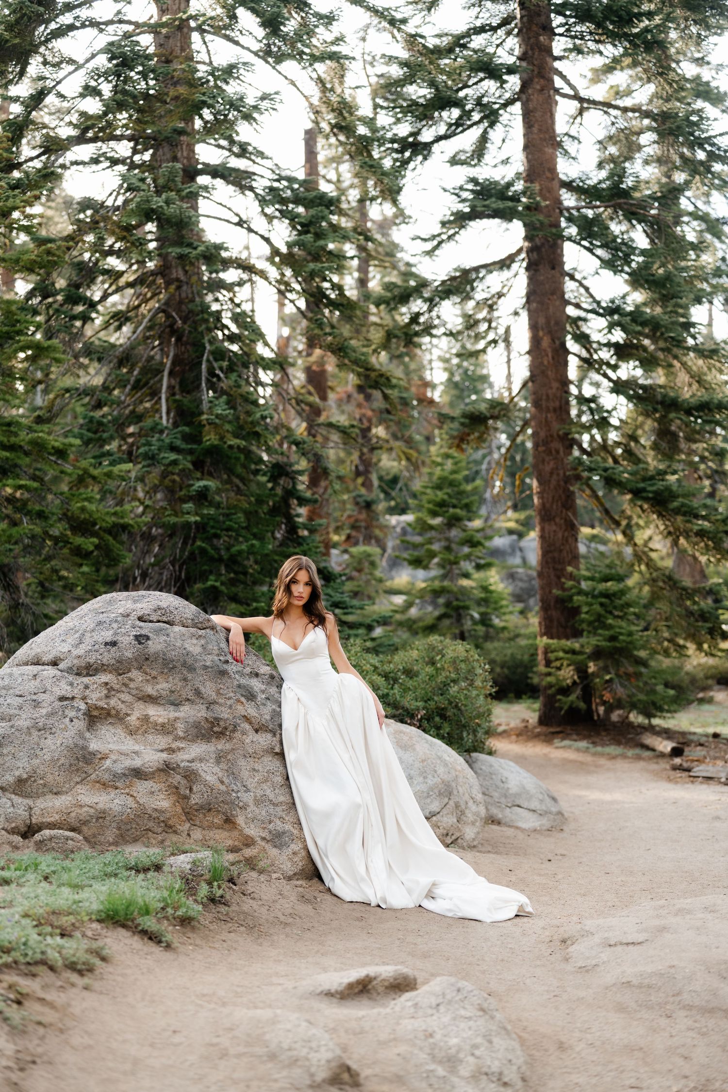 Bride in white flowing gown poses against boulder in pine forest on dirt path.