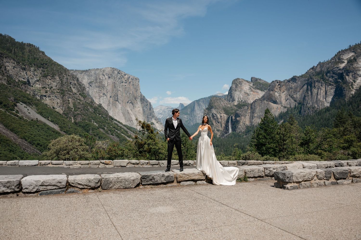 Bride and groom hold hands at Yosemite Valley tunnel view overlook with El Capitan and Bridalveil Fall backdrop.