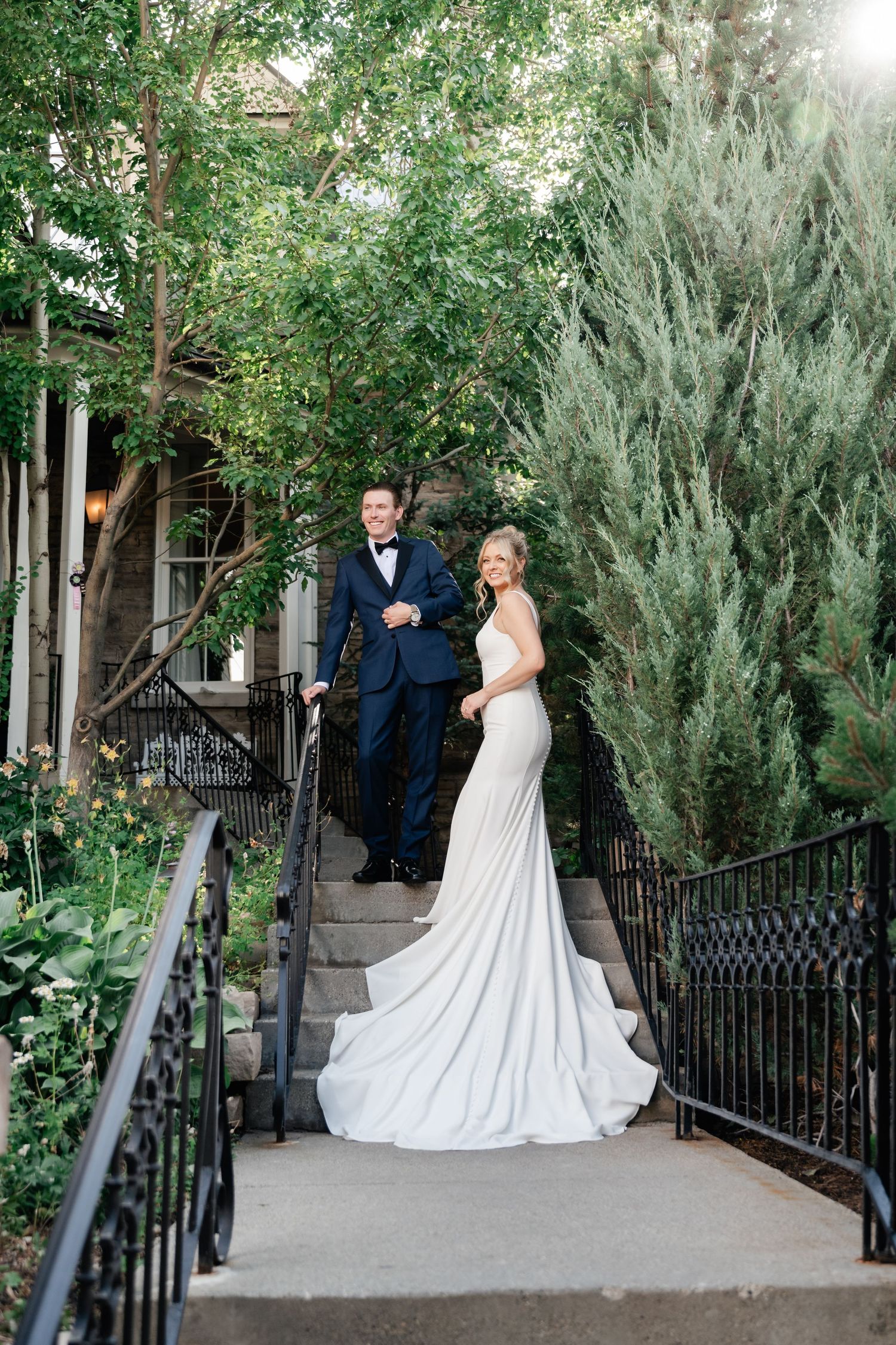 Elegant wedding couple poses along a stone walkway with wrought iron railings and lush greenery.