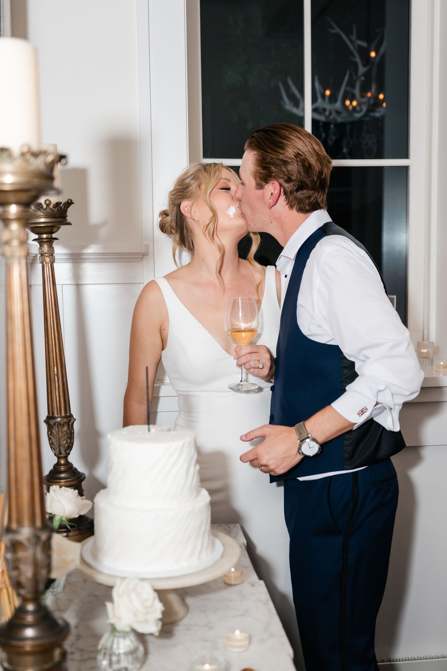 Couple sharing a romantic kiss beside their elegant white wedding cake in an ornate indoor setting.